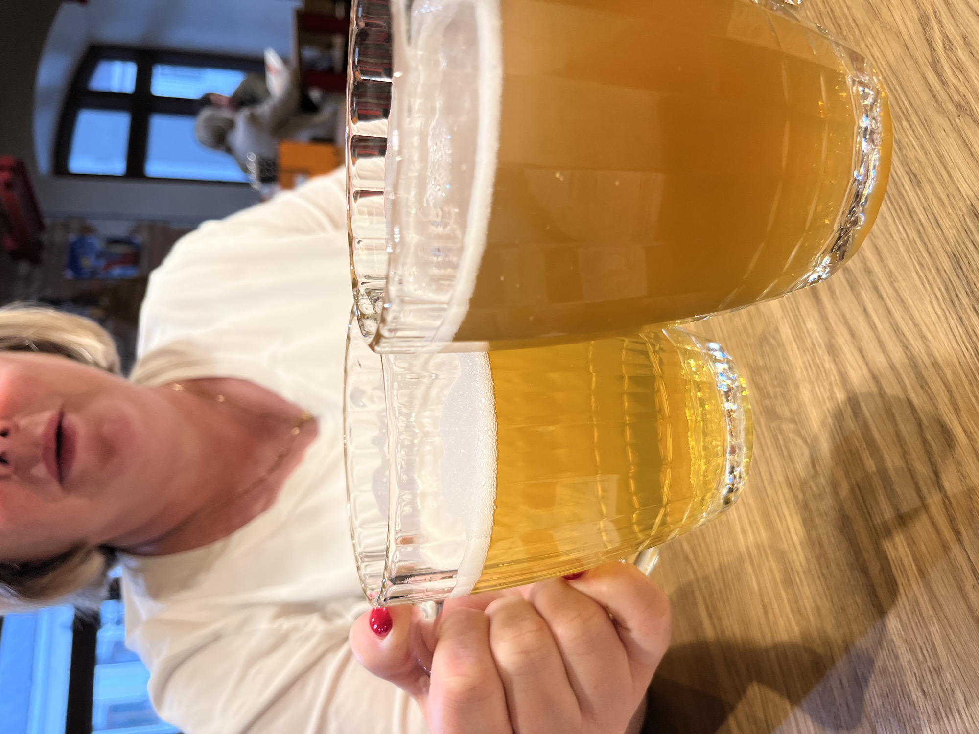 In a close-up shot taken at a low angle, two glasses of beer are prominently displayed on a wooden table. The person holding the left glass, whose hand is visible with red nail polish, is a woman with blonde hair and fair skin, wearing a white top. The background is blurred, showing indistinct figures of other patrons and the interior of a establishment. The scene suggests a casual dining or drinking setting. The specific location in the city of Novo-Nikolskoye, Russia, is not discernible from the visual information provided.