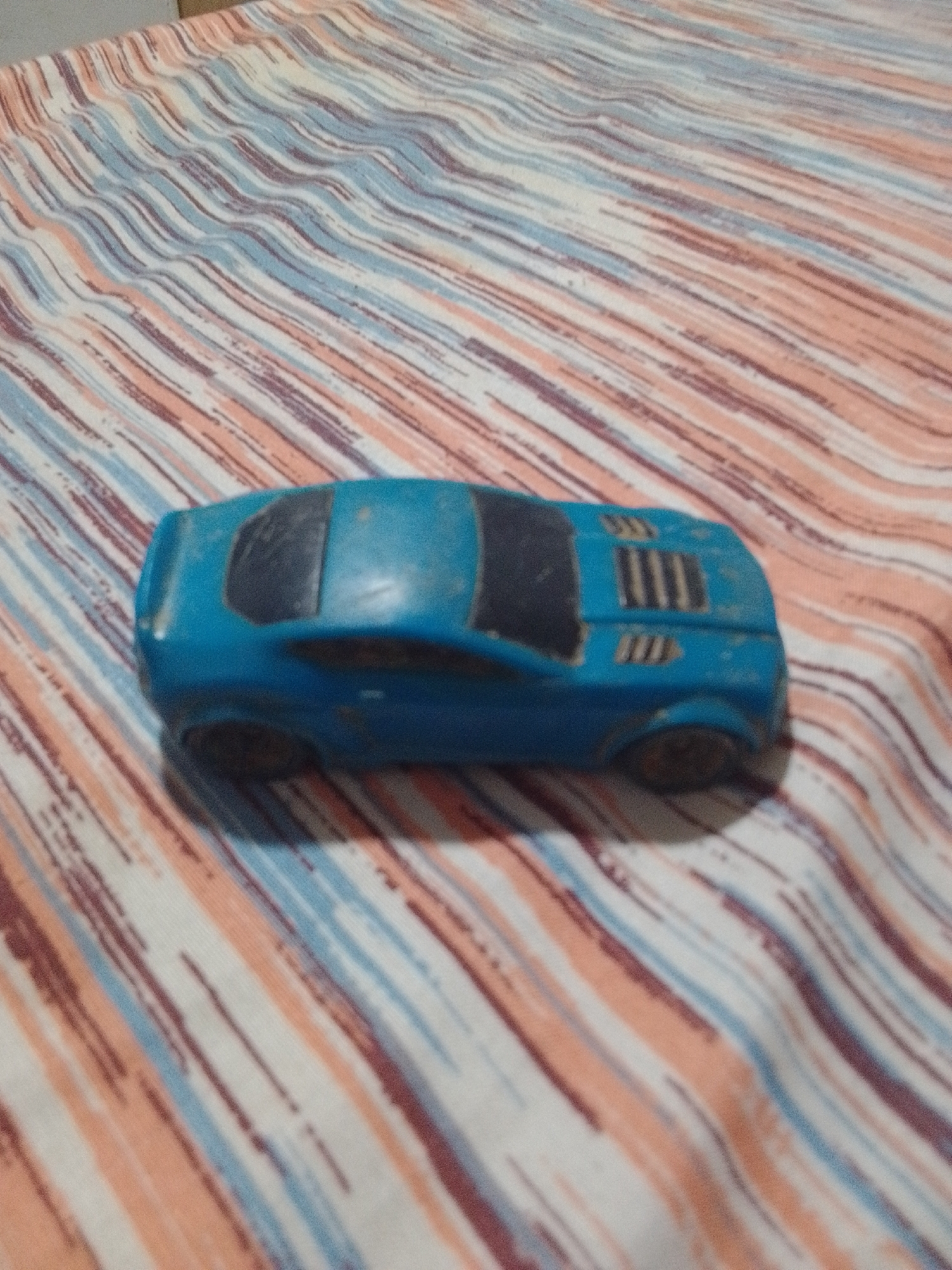 A blue toy car is positioned on a striped fabric surface. The fabric has diagonal stripes in shades of peach, light blue, and dark red. The car appears to be a sports car model, with dark tinted windows and raised elements on the rear. The image is taken from an elevated perspective, looking down at the car. There is no indication of a specific location in La Tagua, Venezuela.