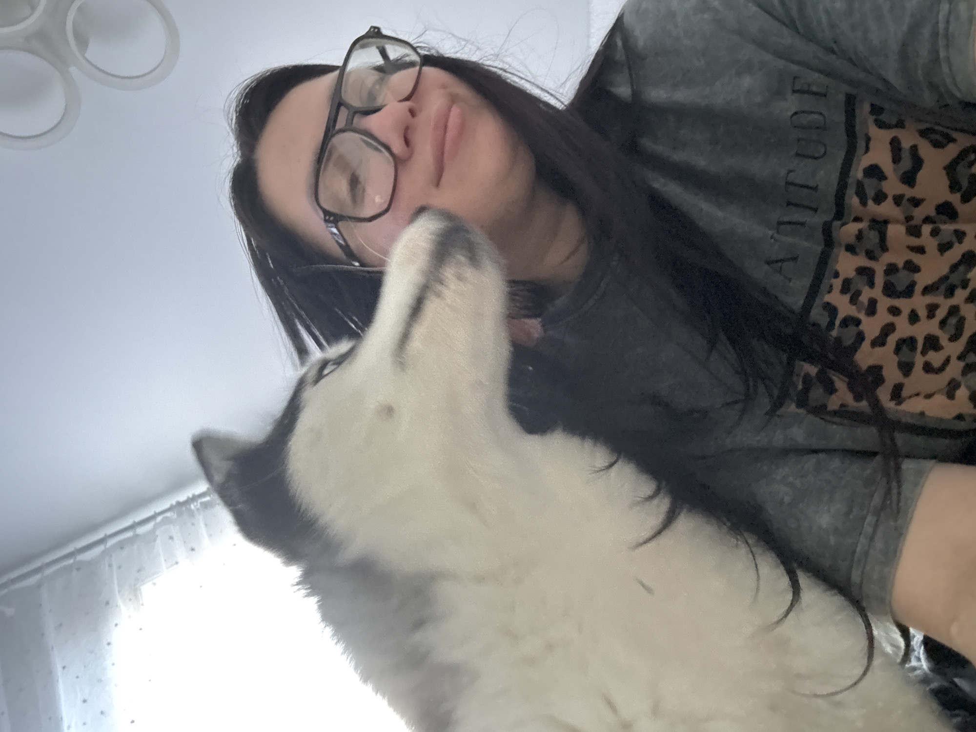 A low-angle shot shows a woman with dark hair and glasses looking downward with pursed lips, a husky dog partially in the frame to her left. The woman wears a grey shirt with "ATTITUDE" printed vertically, alongside a leopard print design. The location appears to be indoors. The city and location are unknown.