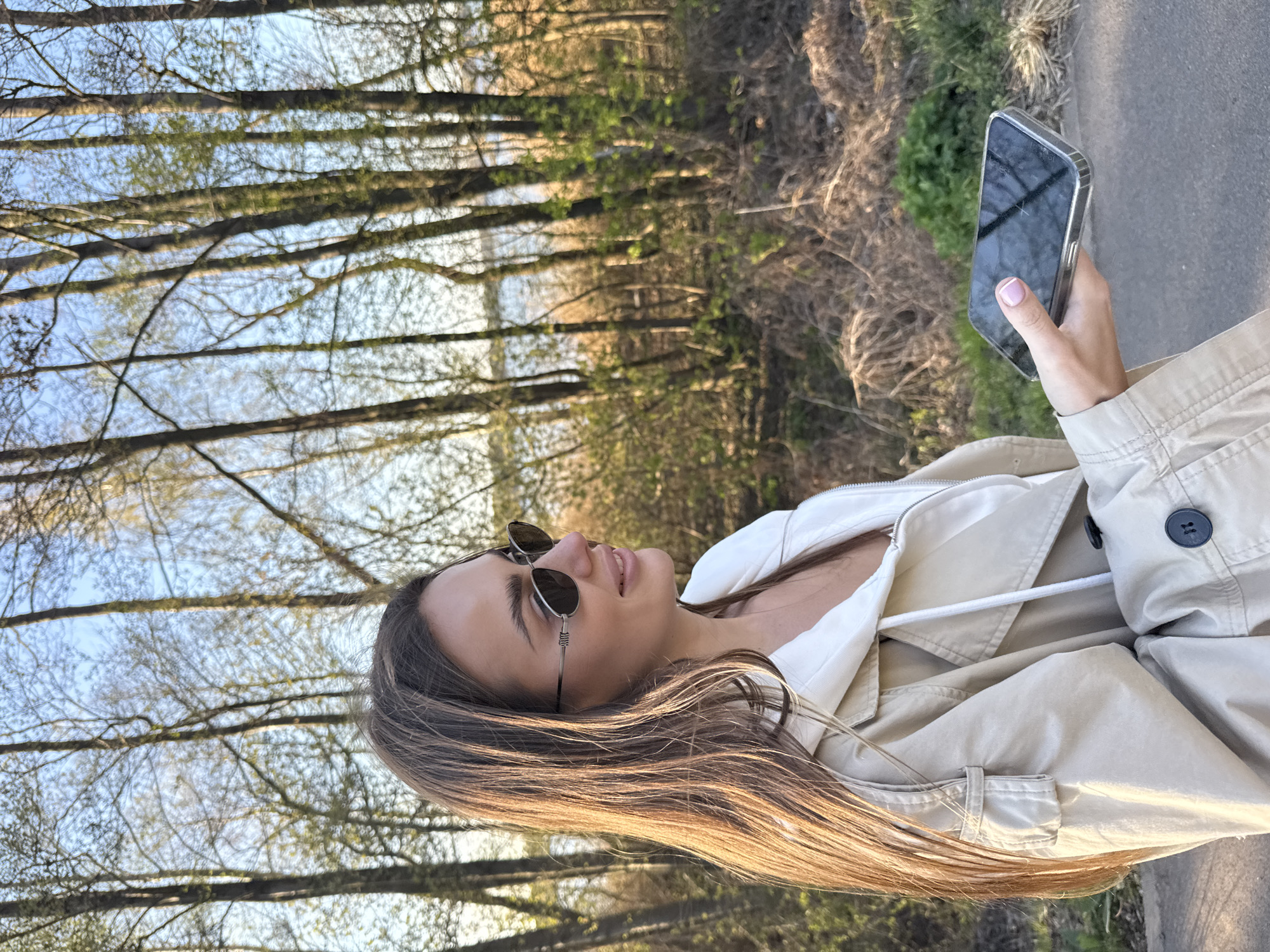 A young woman is depicted outdoors in what appears to be late winter or early spring in Białobrzegi, Poland. She is seen in profile, looking upwards and slightly to her left, with her head tilted back. She wears a light-colored (beige or tan) jacket or trench coat over a white top, featuring a visible drawstring. Round, dark-lensed sunglasses with thin frames are perched on her nose.
Her long, dark brown hair, highlighted by warm, golden sunlight, falls past her shoulders. She holds a dark-colored smartphone in her right hand, encased in a clear protective cover. Her fingernails have a light pink polish. The phone's screen reflects the surroundings, making any text illegible.
The background consists of a natural landscape dominated by numerous tall, deciduous trees, many still bare or just beginning to show small green leaves. A clear, light blue sky is visible through the branches. A paved or gravel path runs to the woman's right, while a body of water, likely a river or lake, is faintly discernible behind the trees on her left, consistent with the Pilica River near Białobrzegi. The lighting suggests early morning or late afternoon, creating a serene and contemplative atmosphere.