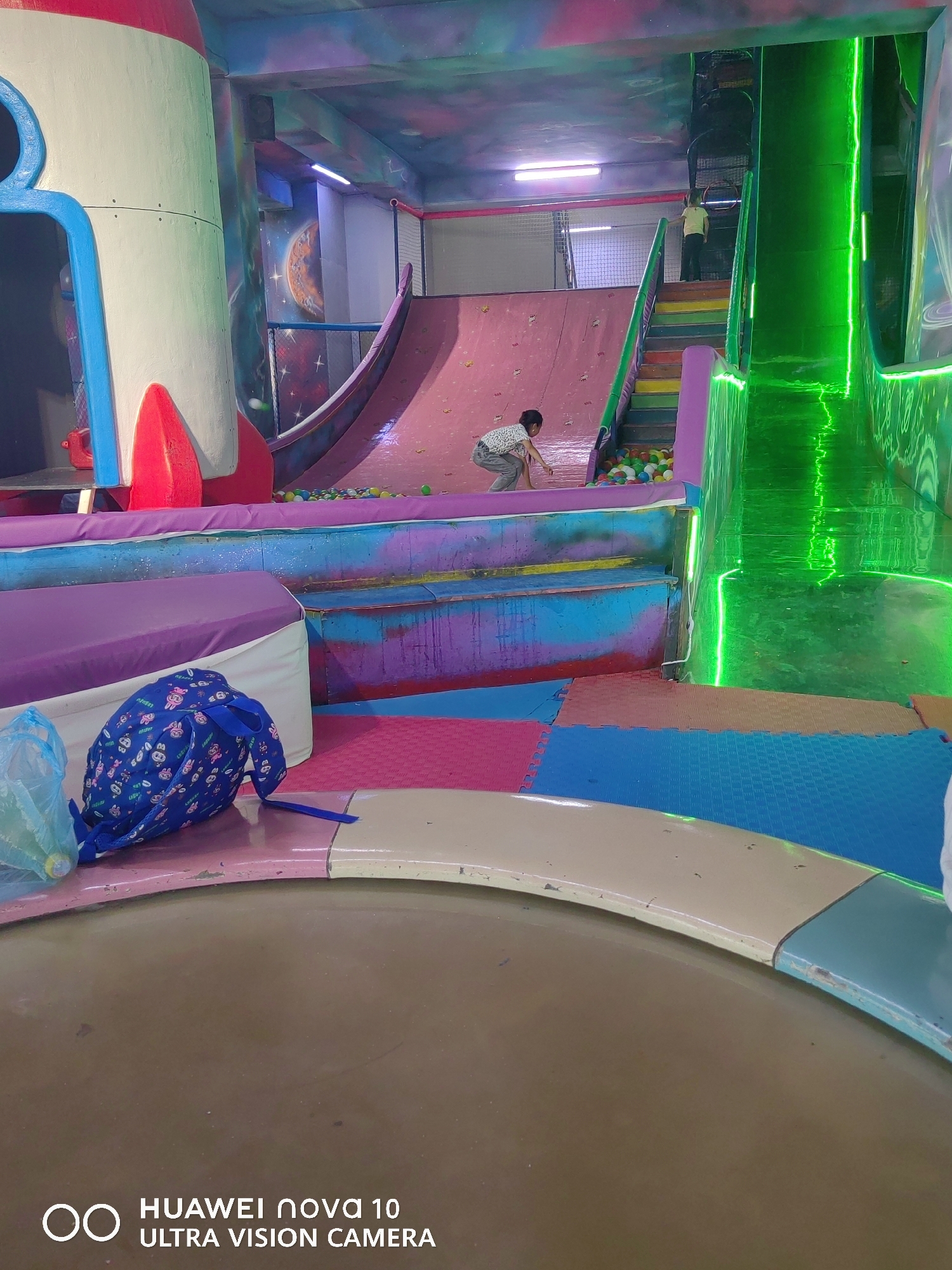 This image shows an indoor playground with a rocket-themed play structure. A young child is visible playing on a pink slide with a ball pit at the bottom. The playground features colorful mats on the floor, a large green slide illuminated by neon lights, and a climbing wall. A blue backpack with a cartoon print is placed on the floor in the foreground. The scene is likely located in an entertainment center, possibly within the city of Özgön, Kyrgyzstan, given the watermark "HUAWEI nova 10 ULTRA VISION CAMERA".