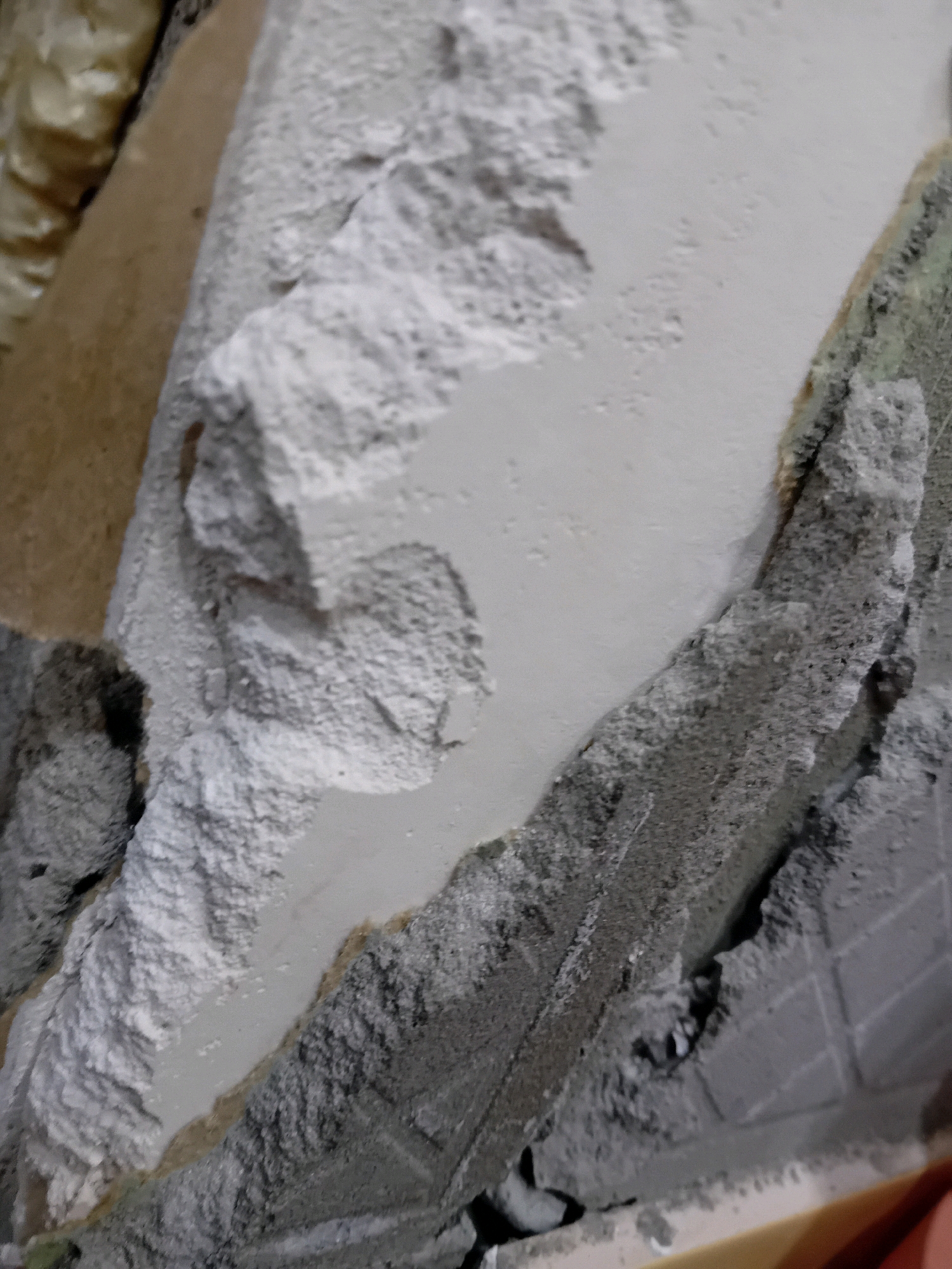 The image shows a close-up, macro view of damaged building materials. The scene is forensic and matter-of-fact, with no people or discernible actions. The environment appears to be a construction or demolition site, with various textures and layers of building materials visible.
In the foreground, there are rough, broken pieces of what appear to be concrete or plaster, with a white powdery substance coating some surfaces. There are also areas of grey, granular material, possibly mortar or aggregate. A tan-colored material, possibly wood or insulation, is visible in the upper left portion of the image, partially covered by the white substance. The background shows a lighter, smoother surface, possibly a wall or ceiling, with some textured areas that might be tiles or another building element.
The scene is devoid of any specific landmarks or features that would allow for definitive identification of its location within Chuhuiv, Ukraine. The image solely focuses on the damaged materials, creating an abstract composition of textures and forms.