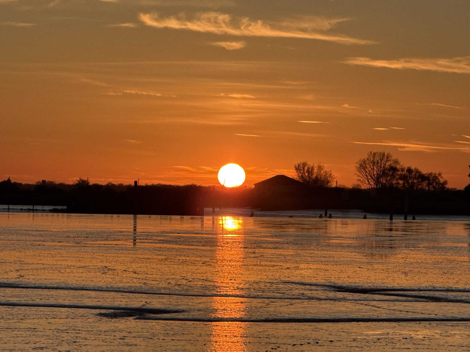 The image shows a sunset over water, likely a broad or river, near Great Yarmouth, United Kingdom. The sun is a bright orb partially obscured by the horizon, casting a distinct, shimmering reflection on the water's surface. The sky is filled with hues of orange and yellow, transitioning to a lighter tone higher up. Thin, streaky clouds are visible in the upper portion of the image. A dark, silhouetted shoreline runs along the midground, featuring a mix of trees and a structure that could be a house or shed. A few posts or pylons are seen in the water closer to the shore. The foreground is composed of shallow water or mudflats, possibly tidal, with subtle wave-like patterns visible. The overall scene conveys a sense of tranquility and a natural landscape.