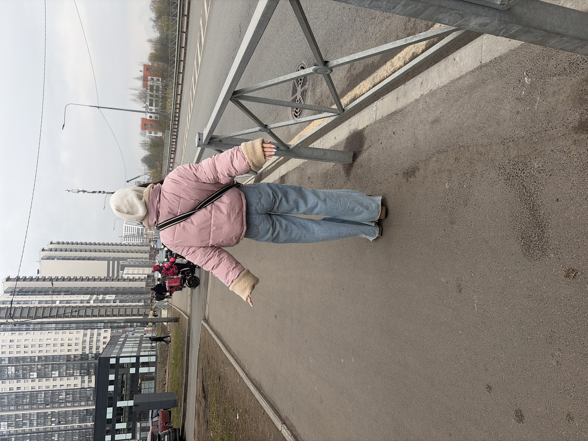 A full-body shot shows a woman with her back to the camera standing on a sidewalk. The woman is wearing a pink puffer jacket with fur-lined cuffs, blue jeans, and a cream-colored hat, and she is carrying a black and white striped bag over her shoulder. She holds a cigarette in her left hand. She is standing next to a metal guardrail that runs along the sidewalk. In the background are tall apartment buildings, and delivery drivers are visible in the distance. The photo was taken in Saint Petersburg, Russia.
