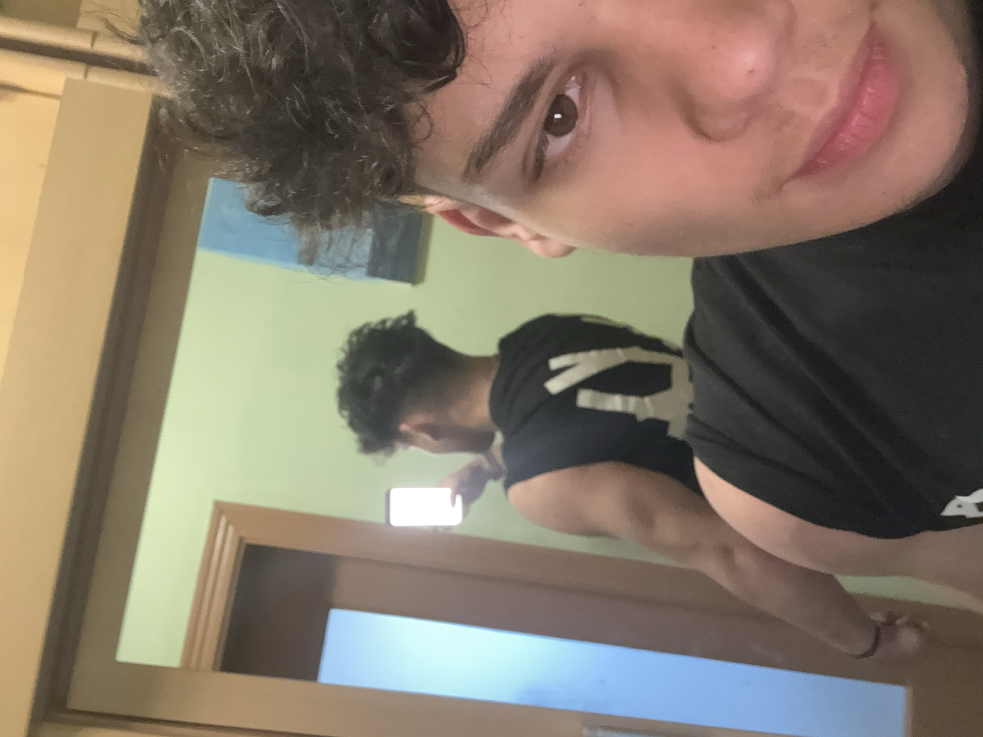 A male person is taking a selfie in front of a mirror. The reflection in the mirror shows the male holding a phone, wearing a black t-shirt, and standing in what appears to be a bathroom or similar space with a light-colored wall. The male's reflection shows their back and upper arm. The immediate environment contains a doorframe and a blurred, light-colored object, possibly a window. The location within Sofia, Bulgaria, is not determinable from the visual information.