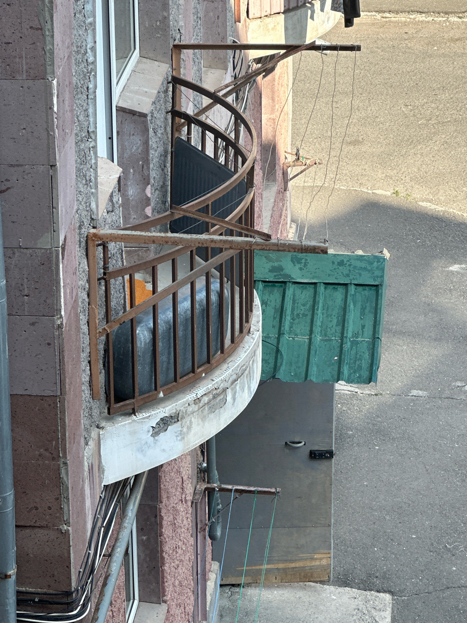 This image depicts a weathered balcony on the side of a multi-story building in the city of Gyumri, Armenia. The balcony features a rusty metal railing with a curved design. Inside the balcony, a large, dark rectangular object, possibly a water tank or storage container, is visible, along with a piece of orange material. A makeshift green corrugated metal awning extends from the railing. The building's facade is constructed with pinkish-brown stone blocks and textured plaster. Below the balcony, a grey metal door with a handle and what appears to be an intercom system is set into the building. Electrical conduits and wires are bundled and run along the exterior wall near the base of the balcony. The ground level shows an asphalt surface and a concrete curb. Several thin, taut lines, likely for drying clothes, are strung from the building outwards. The overall scene suggests a utilitarian, lived-in urban environment with signs of age and repair.