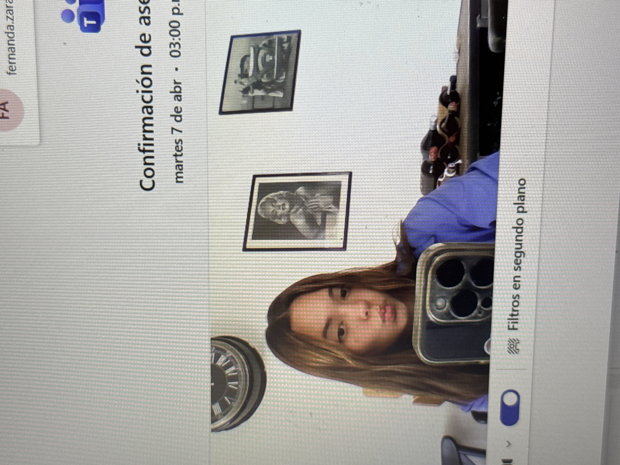 Here is a forensic description of the image:
The image appears to be a screenshot of a computer screen showing a video call or meeting platform, possibly Microsoft Teams. In the upper left corner, there is a user identified as "fernanda.zara" with a "FA" initial icon. Below that, there is a notification stating "Confirmación de ase" followed by the date and time "martes 7 de abr. 03:00 p.m." Two framed pictures can be seen hanging on the wall behind the person in the video.
The main subject of the image is a person facing the camera and holding a smartphone, possibly to record the screen. The phone case is black. The person is sitting on a chair, wearing a purple top, and has light brown hair. A clock is affixed to the wall on the left side of the image.
On the right side of the screen is a portion of a computer program window with the text "Filtros en segundo plano" indicating potential video filter options. There's also an indiscernible object near the upper-right corner, above the individual.
Based on the language, the location might be in a Spanish-speaking country. However, the image itself does not provide any indication of being in San Bernardino Tlaxcalancingo, Mexico, or any specific location.