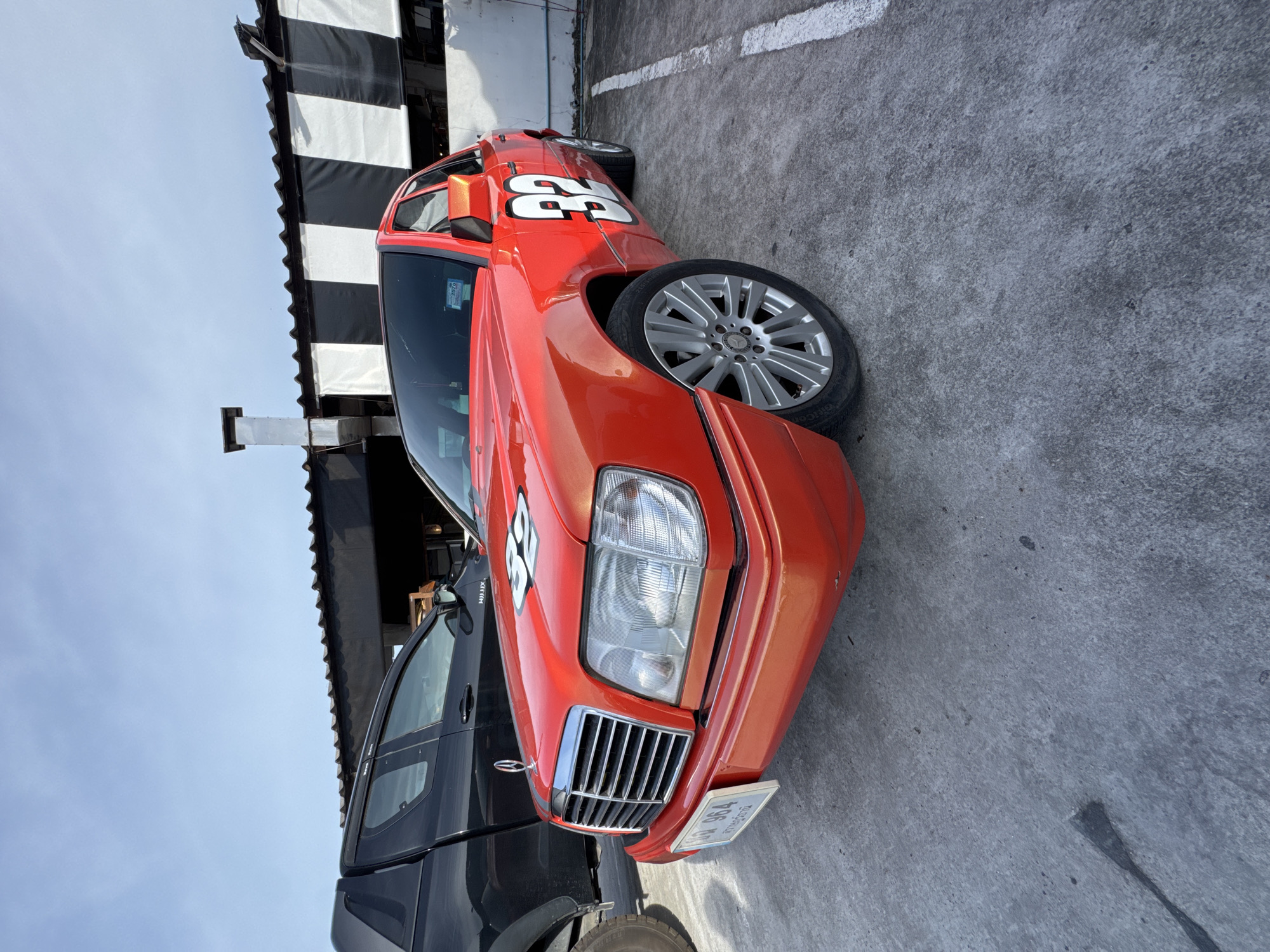 A bright orange Mercedes-Benz sedan with racing numbers "32" on the hood and door is parked on a concrete lot. The car has a chrome grille and silver multi-spoke rims. A black pickup truck is parked to the left of the sedan. In the background, a building with a black and white striped awning is visible. The sky is clear and blue. The scene appears to be in a parking area in ตำบลเกาะพะงัน, Thailand, likely near a commercial establishment.