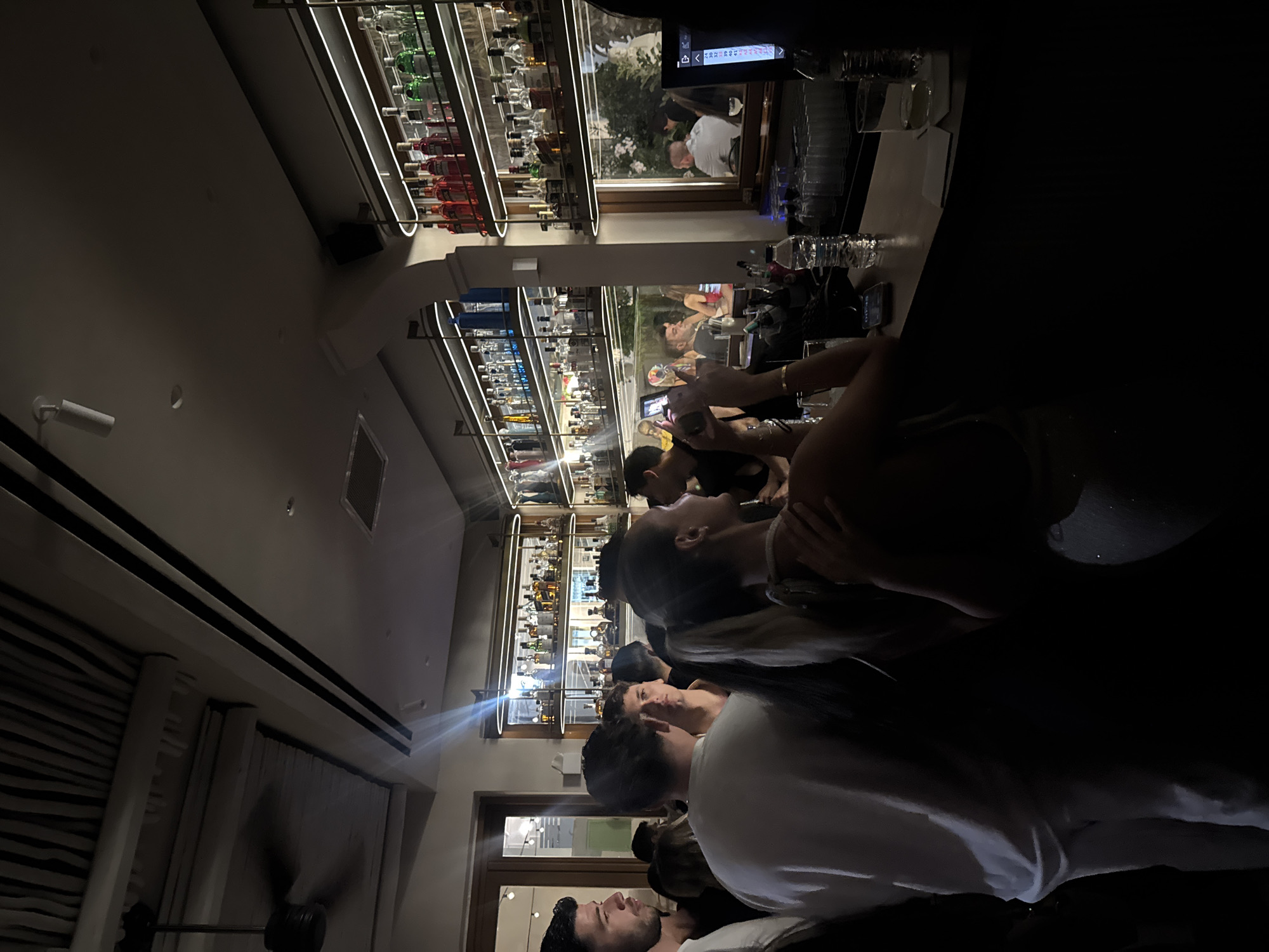 The image depicts an indoor scene at a bar in Naousa, Greece, characterized by dim lighting and a crowded atmosphere. Several individuals are present, with some gathered around a bar area. In the foreground, a woman with her back to the camera, wearing a light-colored top, is looking towards the bar and holding her phone up, possibly to take a photo or video. To her left, a man in a white t-shirt is also looking in the same direction. Behind them, a man with dark hair is visible. The bar itself is stocked with numerous bottles of alcohol on illuminated shelves, suggesting a well-appointed establishment. There are glasses and a bottle of water on the bar counter. In the background, the reflections in a mirror show other patrons and an outdoor scene with some greenery. The ceiling features a textured, possibly wooden, design and a ceiling fan. The overall impression is of a lively evening at a bar or nightclub.