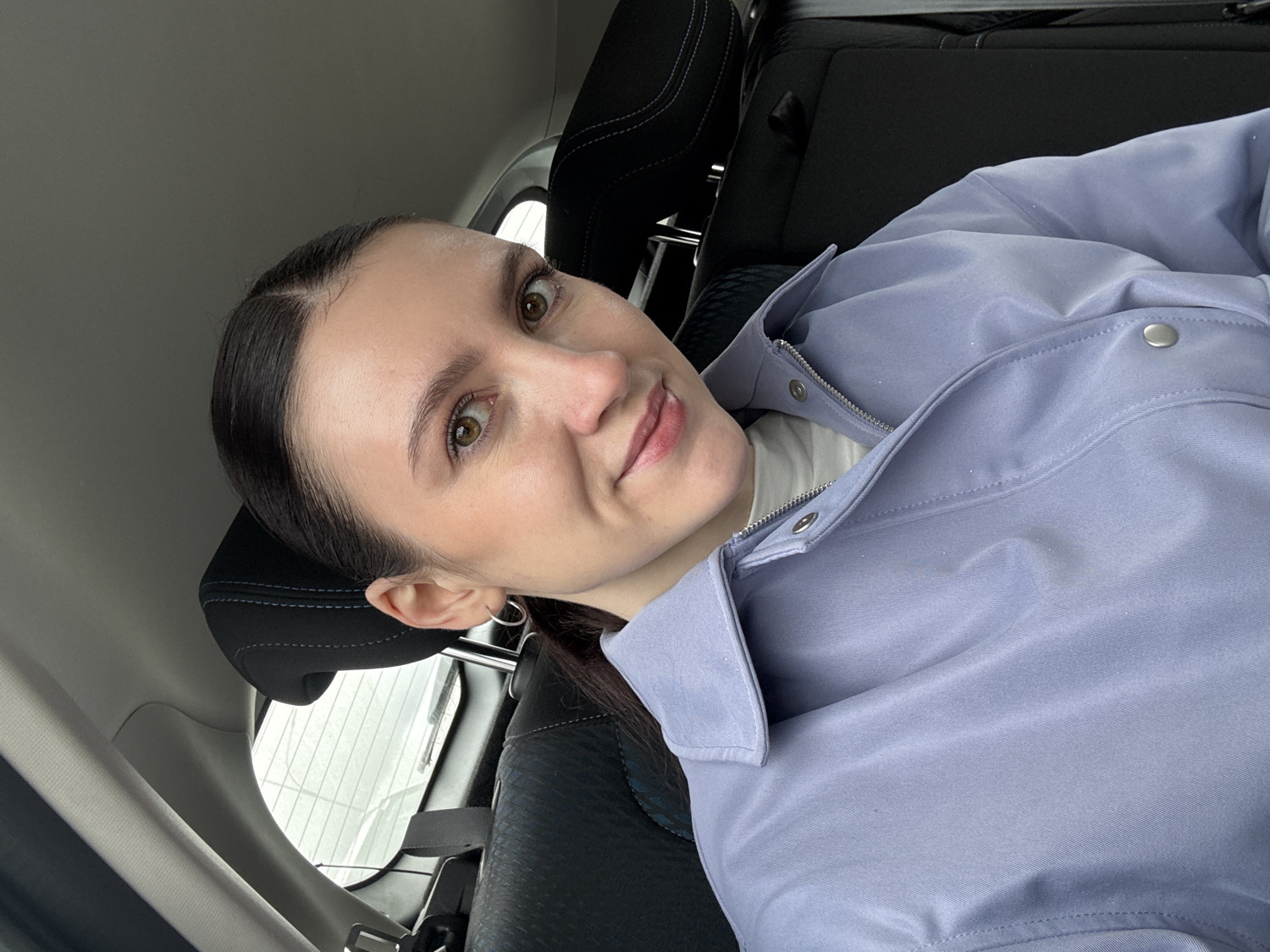 A young woman is depicted in a selfie-style portrait, positioned inside what appears to be a car. She has medium skin, dark brown hair parted on the side and pulled back, and brown eyes that look directly at the camera. A slight, gentle smile plays on her lips. She is wearing a light lavender or periwinkle-colored jacket with silver snap buttons and a zipper, over a white top, possibly a turtleneck. A small silver hoop earring is visible on her left ear.
The setting is the interior of a vehicle, suggested by the black car seats and headrests visible behind and around her; one headrest features white stitching. The car's ceiling or headliner is a light grey. Through a window visible behind the headrest, a blurred, light-colored outdoor scene can be discerned, consistent with an overcast sky or possibly a snowy environment. A seatbelt mechanism is also visible on the left side of the frame. The soft, diffuse lighting suggests it is daytime, likely under overcast conditions. There is no visible text in the image. The woman's direct gaze and subtle smile indicate she is posing for the photograph. The general location context is Kazan, Russia.