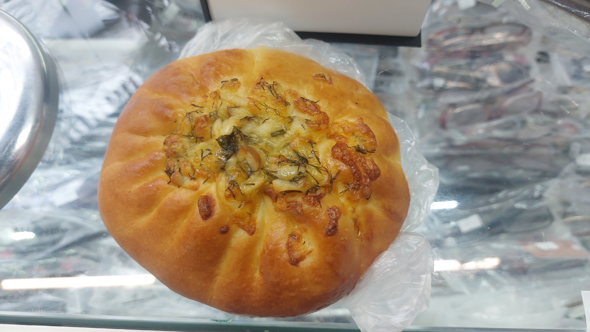 A top-down, eye-level shot depicts a round, golden-brown baked pastry in a clear plastic bag, resting on a glass surface. The pastry is topped with melted cheese and green herbs. The background includes out-of-focus items behind the glass, including what appear to be eyeglasses. The scene is located in Novosibirsk, Russia.