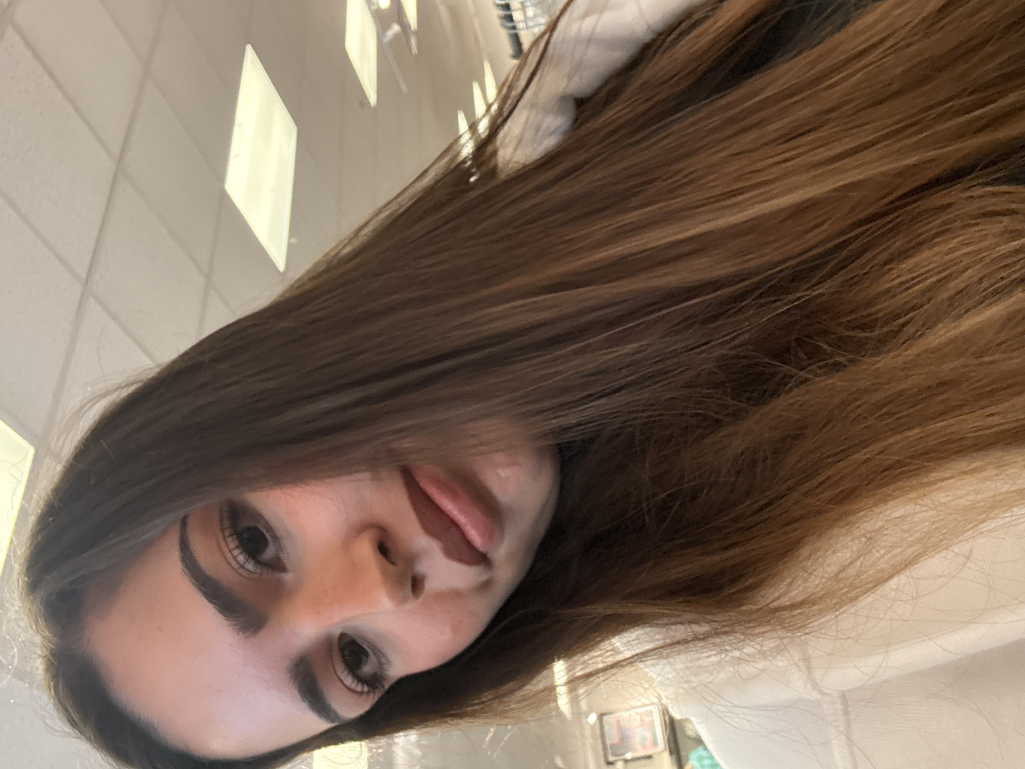 A close-up, angled selfie captures a young woman with long, light brown hair partially obscuring her face. Her dark eyebrows frame brown eyes, and freckles are visible on her fair skin. She has full lips. The background shows a white tiled ceiling with recessed lighting, indicating an interior space. The photo appears to have been taken indoors, possibly in a public building. San Marcos, United States of America cannot be determined from the image.