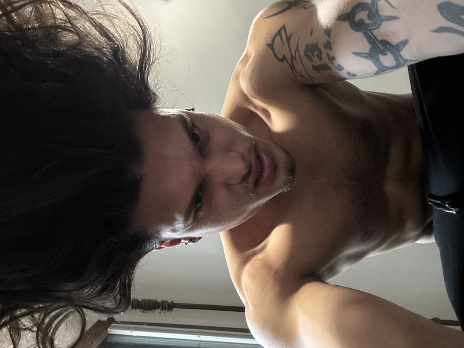 The scene is located indoors in the city of Sofia, Bulgaria. A male individual, shirtless and appearing to be in his twenties or early thirties, is captured in a close-up, low-angle perspective. His dark, voluminous hair stands dramatically upright, extending beyond the top of the frame. He has an intense, snarling facial expression, with his mouth slightly open, lips pulled back, and brows furrowed. Dark eyes gaze directly forward. Small hoop earrings are visible in his left ear and a stud in his right. His muscular torso is bare, showing defined shoulders, chest, and abdominal muscles. On his right upper arm, multiple dark tattoos are visible: a large, stylized crown-like or tribal design, the number "13," and Cyrillic script which appears to be "БЪЛГАРИЯ." Below these, on his bicep/forearm, is a barbed wire or chain-link pattern. His left arm extends towards the camera, suggesting a selfie. The lower portion of the image shows black athletic pants or shorts with a visible drawstring. The background consists of a plain, light-colored wall, with a blurred vertical element, possibly a window frame or furniture, visible on the far left, suggesting ambient light source.