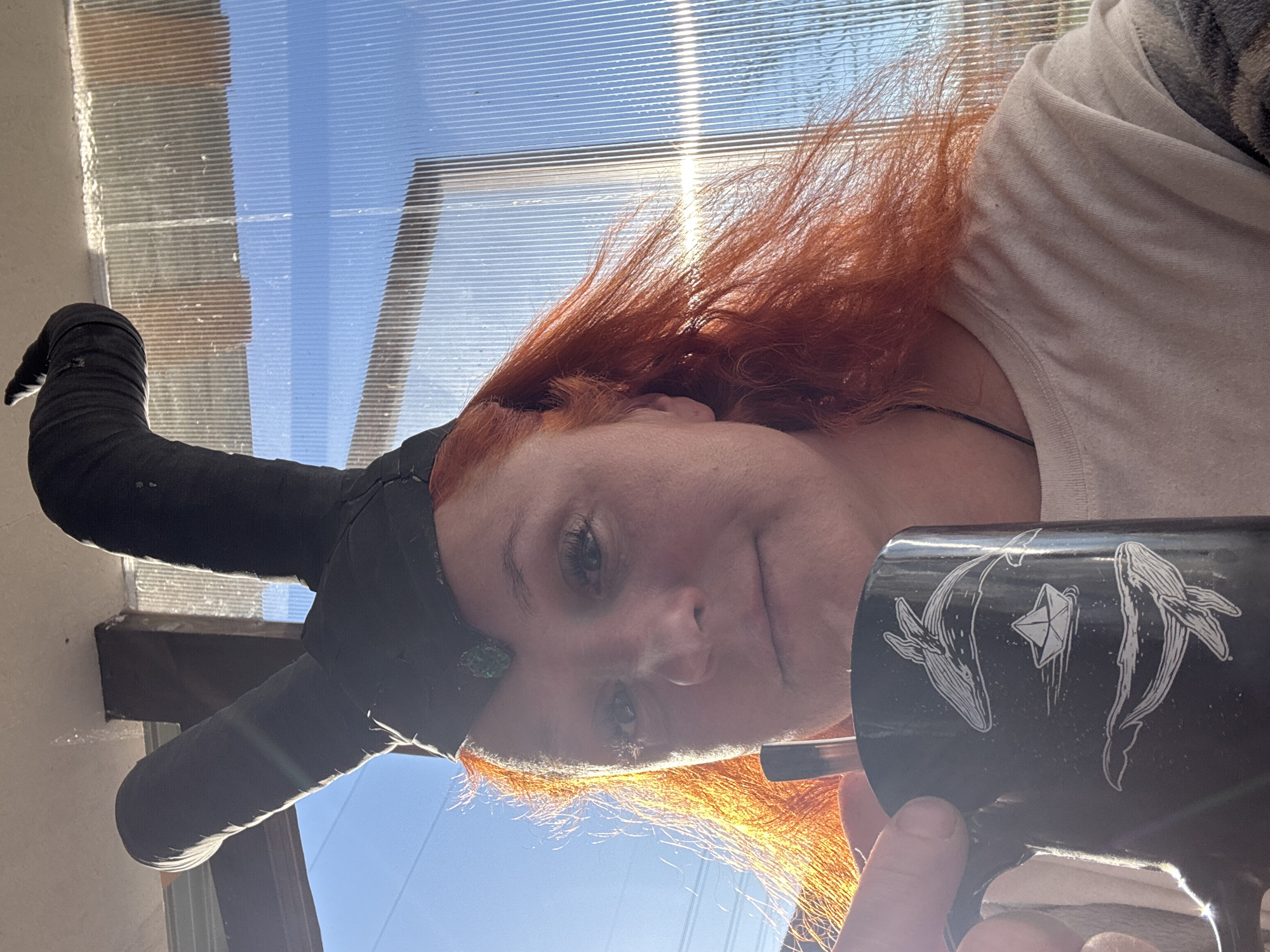 A person with reddish hair and a light complexion is holding a black mug with white whale designs. The person is wearing a light-colored t-shirt and a black head covering. In the background, there is a window with a view of a bright blue sky. The photograph appears to be taken indoors, with sunlight streaming in. The image does not provide enough information to verify the location in the city of Chaikovsky, Russia.