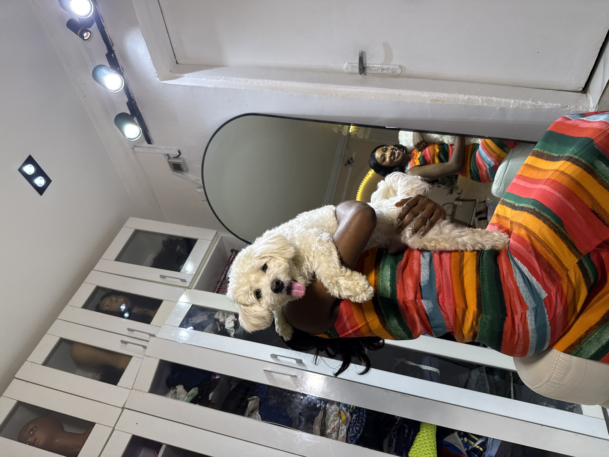 A medium shot shows a Black woman holding a small, fluffy white dog in an indoor environment that appears to be a closet or dressing area. The woman, wearing a sleeveless, vibrantly striped dress with colors including red, orange, yellow, green, and blue, is facing the viewer and holding the dog up towards the camera. The dog has its tongue slightly out and appears to be looking at the camera. Behind the woman, a large arched mirror reflects the woman and the dog, as well as a white door with a silver handle to the right of the mirror. To the left of the woman and the mirror is a tall white wardrobe with glass-fronted doors, revealing clothing and mannequins with wigs inside. The ceiling is visible at the top of the image, featuring track lighting with multiple spotlights and a square recessed light fixture with two bulbs. The overall impression is of a private, possibly domestic, space designed for clothing storage and preparation. The setting is in Abuja, Nigeria.