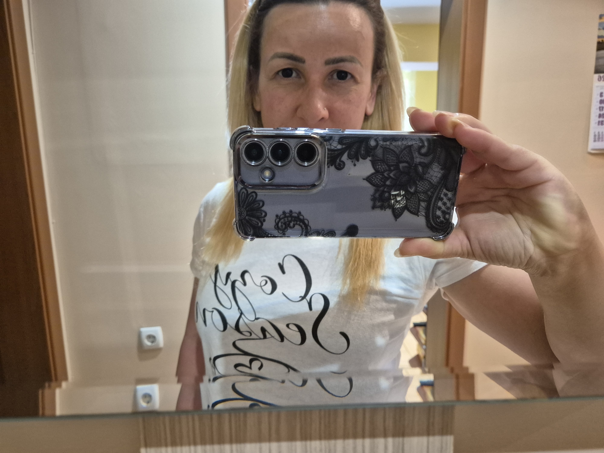 A woman is taking a selfie in a mirror. She is holding a smartphone with a clear case decorated with black lace patterns and a triple camera module. She is wearing a white t-shirt with black calligraphy text. The background appears to be a room with a wall, a doorframe, and two white electrical outlets reflected in the mirror. A calendar is visible on the wall in the upper right corner of the reflection. The lighting is somewhat dim.