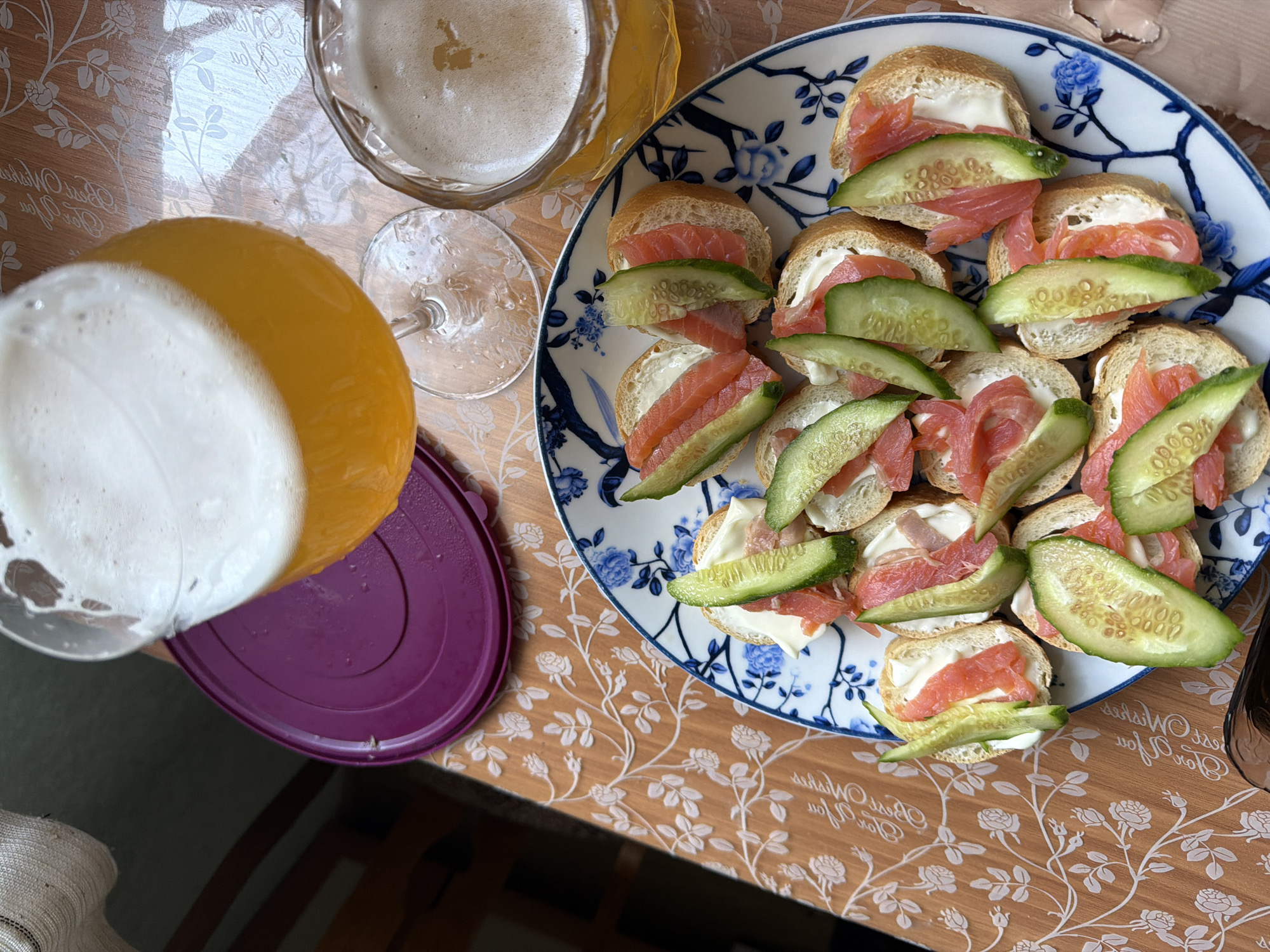 The image depicts a table setting, likely indoors. On the right, there's a decorative plate with small open-faced sandwiches topped with cream cheese, salmon, and cucumber slices. To the left, a pitcher containing an orange-colored liquid with a foamy top is positioned next to an empty wine glass and a partially filled glass of beer. The table is covered with a floral-patterned tablecloth or placemat. Based on the style of decoration and the food items, the location is presumed to be Saint Petersburg, Russia. No people are visible in the frame.