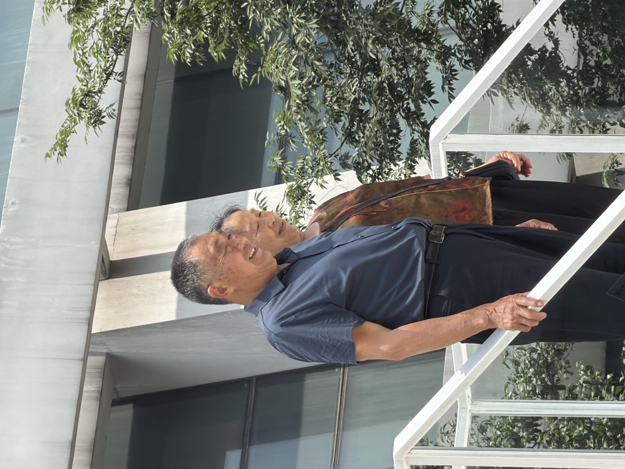 In Yuzhong District, China, an older Asian man and an older Asian woman are depicted walking up a white staircase outdoors. The man, on the left, is wearing a dark blue collared shirt and dark pants. He is smiling and looking to his left. The woman, on the right and slightly behind him, is wearing a patterned top with floral designs and dark pants. She is looking down. Both are on a staircase with white railings and glass panels, with lush green foliage visible to their right and behind them. The background features a modern building with large windows and concrete architectural elements.