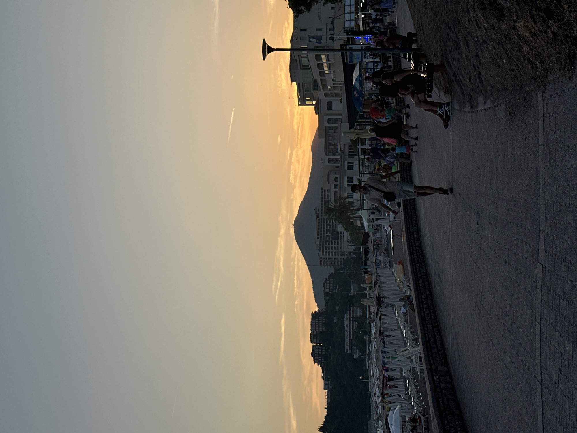 This is a silhouette photograph taken at sunset on a beach promenade in Boreti, Montenegro. The sky is a gradient of pale blue to warm orange, with thin clouds and a faint contrail visible. In the background, a large mountain with a structure on its peak dominates the horizon, flanked by buildings and dense foliage on a hillside. The foreground features a paved walkway with people strolling and sitting. A stone wall separates the walkway from a beach lined with numerous rows of white beach umbrellas and loungers, most of which appear unoccupied. To the right of the walkway, a grassy embankment slopes down, with a dark wooden bench occupied by two individuals. A lamppost stands near the buildings. The overall atmosphere is calm and serene, capturing the end of a day at the seaside.
