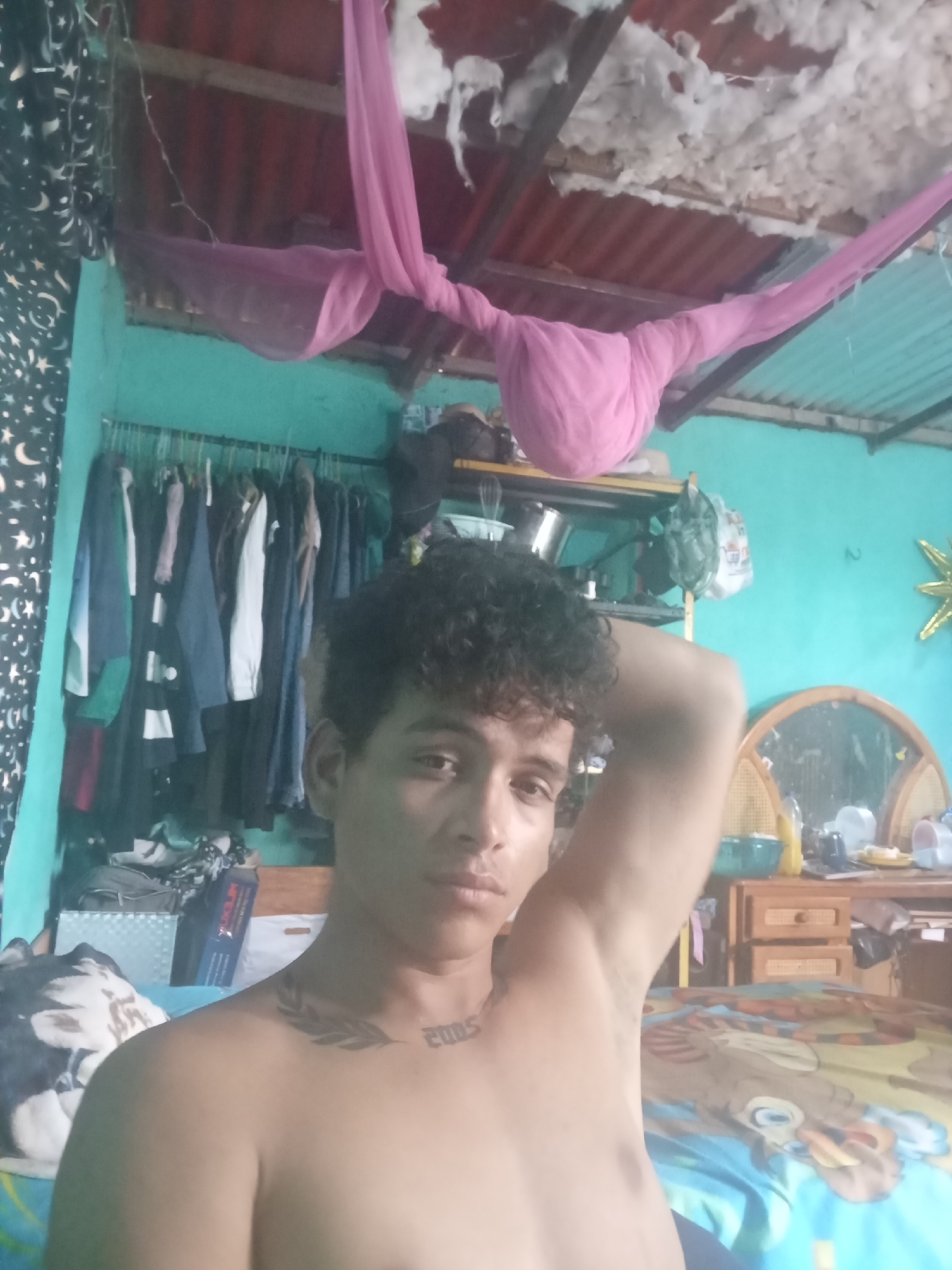 The image shows a young adult male in what appears to be a bedroom. He is shirtless and has curly dark hair. His left arm is raised, resting behind his head. Tattoos are visible on his chest and collarbone. The room has teal-colored walls and a corrugated metal roof. Clothing is hung on a rack on the left side of the image. A pink fabric is draped from the ceiling. Various items are on shelves and a dresser in the background, including a mirror and some bottles. The overall impression is a candid, casual self-portrait taken indoors. The provided hints indicate the location is Altagracia, Venezuela.