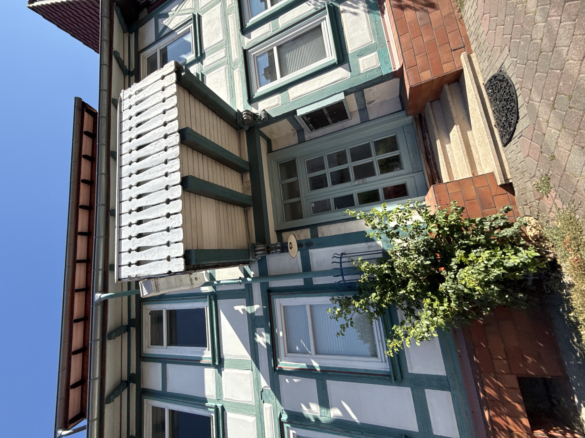 This image shows the exterior of a half-timbered house, likely located in Bad Sachsa, Germany, on a clear, sunny day. The house features white stucco walls with dark teal timber framing. A small balcony with a decorative wooden railing is situated above the entrance. The entrance itself is a light blue door with glass panes, approached by a set of stone steps. A green bush is visible to the left of the entrance. The ground in front of the house is paved with interlocking bricks, and a black doormat is placed on the ground near the steps. The architectural style suggests it could be in a historic district of the town.