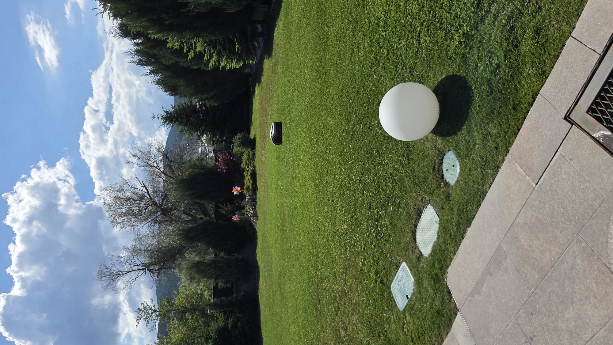 The image shows a well-maintained residential backyard in Bania, Bulgaria. The foreground features a paved patio area and a bright green lawn. Several circular, green access covers are embedded in the lawn. A large, white spherical lamp sits on the grass. Further into the yard, a small robotic lawnmower is visible. In the mid-ground, a decorative pinwheel with colorful blades is planted in a flower bed. The background is dominated by a dense line of evergreen trees, with a larger, bare deciduous tree situated to the left. Beyond the trees, a gently sloping hill with scattered houses and more trees can be seen. The sky is partly cloudy with dramatic white clouds against a blue sky.
