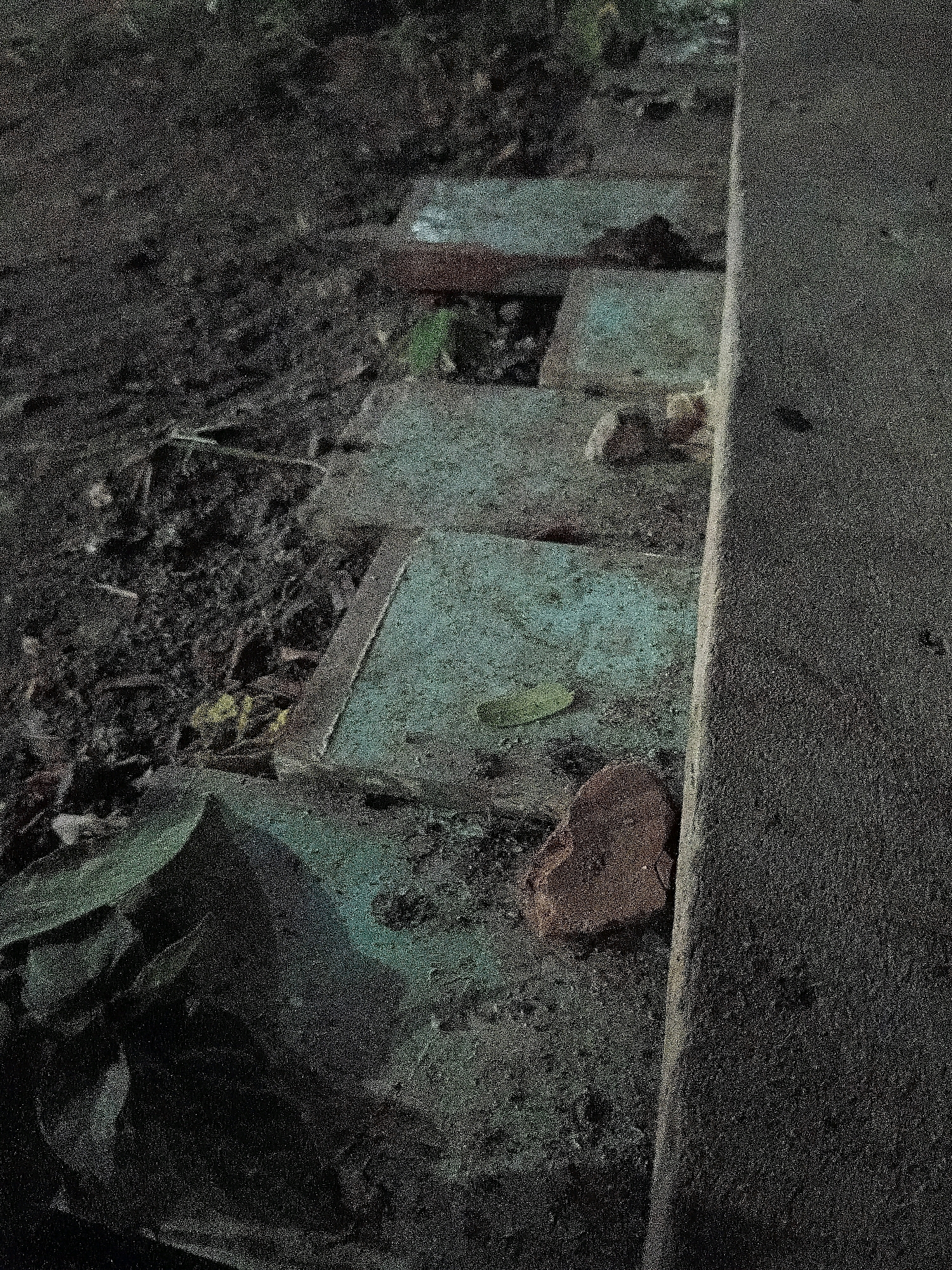 The image shows a close-up, low-angle view of a section of stone steps or paving stones near a concrete curb, likely in an outdoor urban environment. The steps are worn and appear to be made of a light blue-grey material, possibly concrete or a type of tile. They are unevenly laid, with some gaps and debris in between. Loose soil, dirt, leaves, and a broken piece of brick are scattered on and around the steps. A large, dark, irregularly shaped object, possibly a rock or a piece of landscaping, is visible in the lower left corner of the frame. The overall scene is gritty and somewhat neglected, with a focus on the texture and wear of the materials. The lighting is dim, and the image has a grainy quality, suggesting it was taken in low light conditions or with a high ISO setting. There are no people or animals visible in the image, and no specific landmarks or details that would definitively place it in Cikal, Indonesia, or any other specific location.