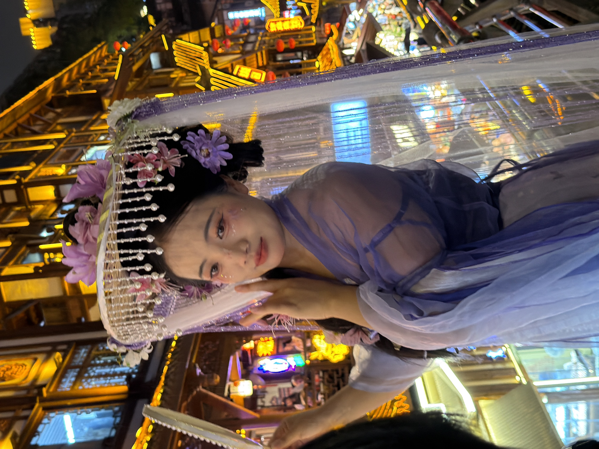 A young woman, adorned in a traditional Chinese costume and ornate headdress featuring flowers and beads, stands in a vibrant, dimly lit streetscape. The background is dominated by multi-story buildings with illuminated windows and glowing signs, suggesting a commercial or entertainment district. The woman's makeup is elaborate, with pink blush and glitter on her cheeks. She is wearing a flowing purple and white garment, possibly made of sheer fabric. Her left arm is bent at the elbow, and her hand is positioned near her face, with her fingers extended. The overall scene depicts a festive or cultural event, likely in a historical area of Yuzhong District, China, given the architectural style and the woman's attire.