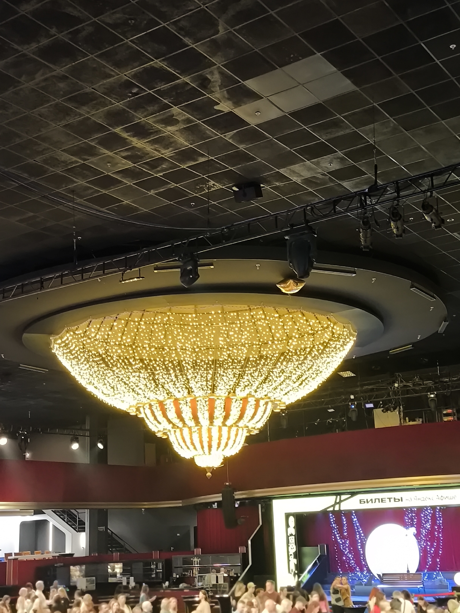 A large, ornate chandelier hangs from a dark, grid-patterned ceiling in a dimly lit venue. The chandelier is circular and covered in numerous small, bright yellow lights, creating a shimmering effect. Below the chandelier, a multi-tiered dark grey structure is visible. Further down, a bright red wall forms a backdrop for a stage area. On the stage, a large screen displays text in Russian, and a bright white circular light illuminates the center. A crowd of people are gathered in the foreground, facing the stage. The scene appears to be an indoor event or performance space.