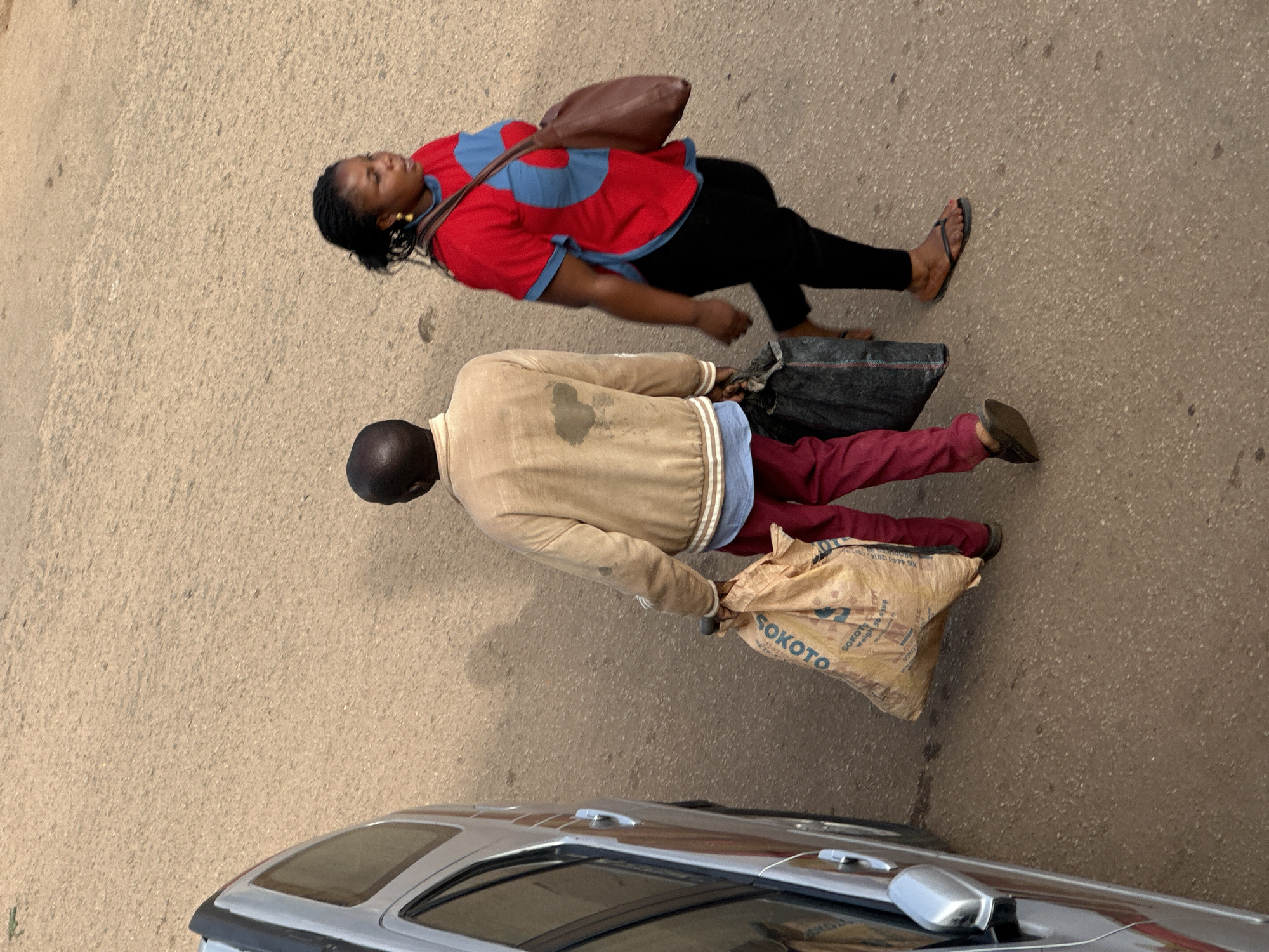 In the city of Bukuru, Nigeria, two individuals are walking on a paved road. The person on the left, identified as a male, is wearing a light-colored jacket over a blue shirt and red pants, carrying a large, beige bag in his left hand and a dark-colored bag in his right. The person on the right, identified as a female, is wearing a red and blue shirt, black pants, and flip-flops. She has a brown shoulder bag and is walking with her right arm slightly raised. A silver vehicle is parked on the left side of the frame. The ground is a textured asphalt surface.
