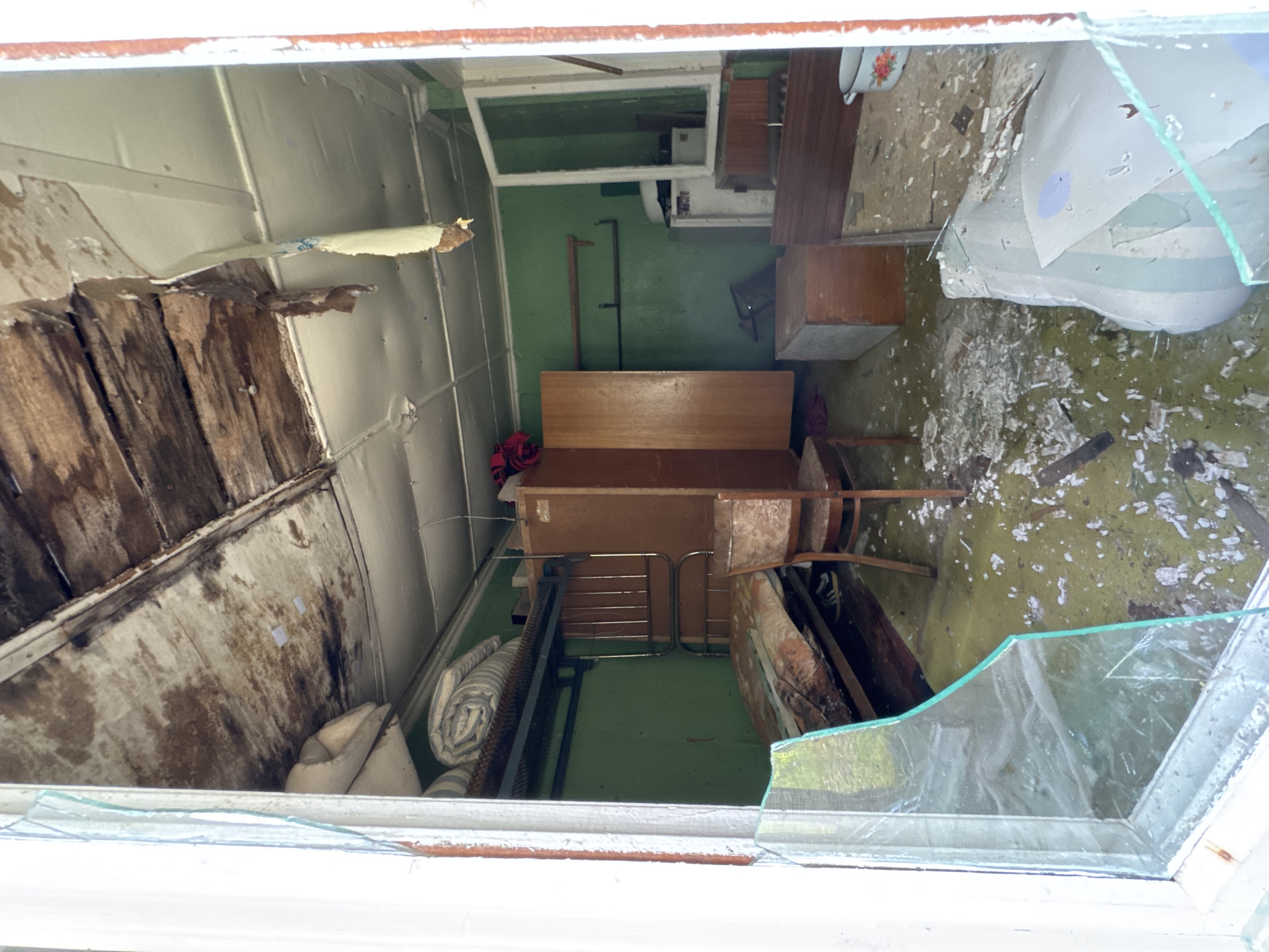 The image shows the interior of a derelict room, likely a bedroom or dormitory, viewed through a broken window. The room is in a state of disrepair, with debris scattered across the floor and water damage evident on the ceiling.
The flooring appears to be a greenish linoleum or painted concrete, covered in dust, broken plaster, and other debris. The walls are painted a dark green color, and in some areas, the paint is peeling. The ceiling is a grid of panels, with significant water stains and sagging. In some sections, the underlying wooden beams are exposed and decaying.
Furniture in the room includes a metal bunk bed with a mattress on the lower level, a wooden wardrobe, a small wooden chair with upholstered seating, and a wooden chest. There are also shelves on the wall, one of which holds a small appliance that resembles a refrigerator or a freezer. A rolled-up mattress or bedding is visible on the top bunk.
The window through which the image is taken is shattered, with large shards of glass lying on the floor inside and outside the frame. The broken glass frames the scene, creating a barrier between the viewer and the interior.
The image does not provide any specific landmarks or contextual clues to pinpoint the exact location within the city of Ikhala, Russia. The overall impression is one of abandonment and decay.