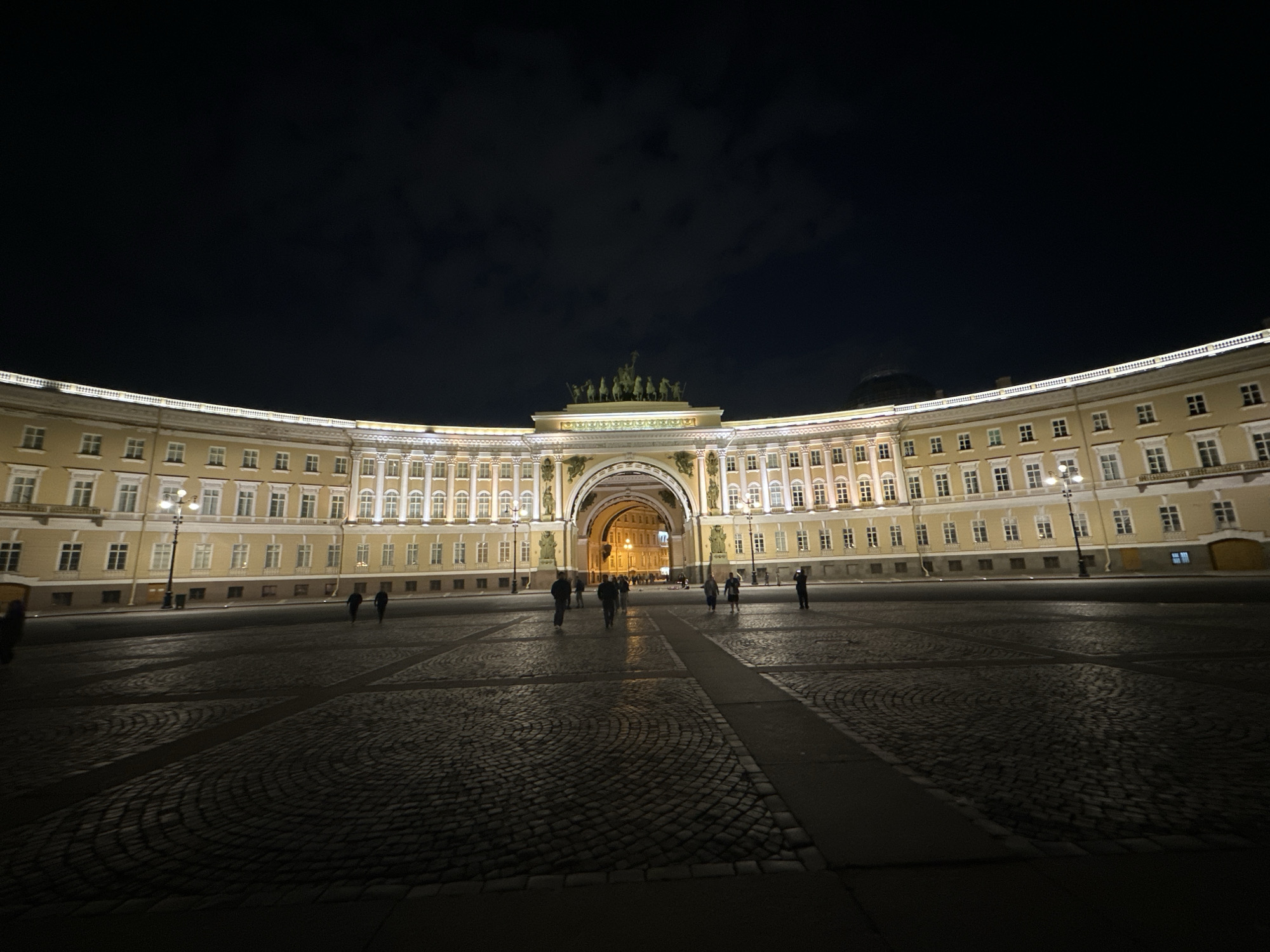 The image shows the Palace Square in Saint Petersburg, Russia, at night. The General Staff Building with its triumphal arch is illuminated and visible at the center of the image. The square itself is paved with cobblestones. Several people are walking across the square, their forms indistinct in the darkness. Street lamps are lit, adding to the illumination of the building. The sky above is dark with clouds.