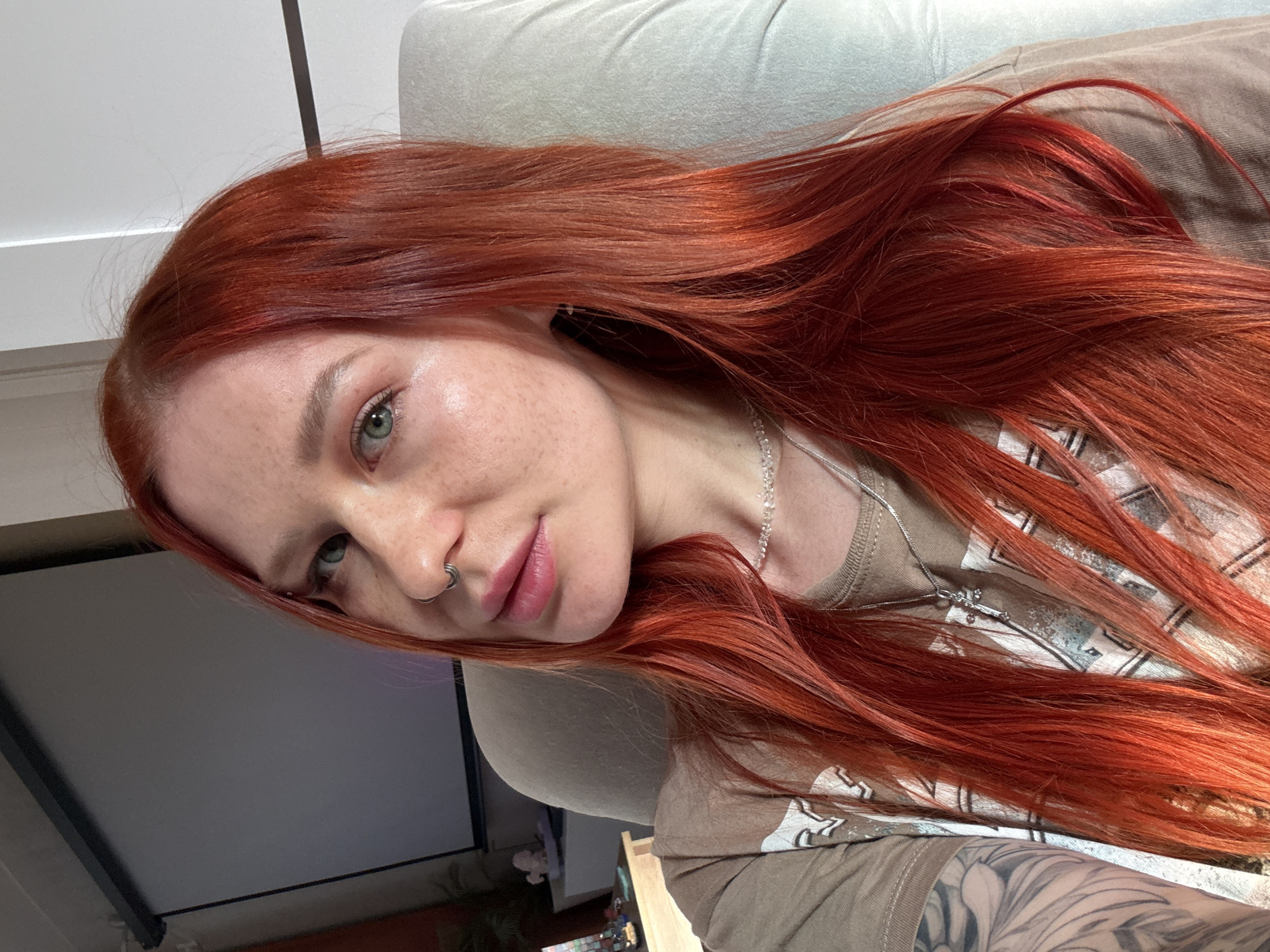A red-haired woman with freckles and a nose ring is lying on a light-colored upholstered piece of furniture, possibly a couch or chair. Her long hair is spread around her on the furniture and over a light brown t-shirt with a design. She is wearing a delicate silver necklace. The woman has green eyes. The location cannot be determined from the image, so it cannot be placed in Rostov-on-Don, Russia. In the background is a gray surface framed in black, possibly a television screen or window.