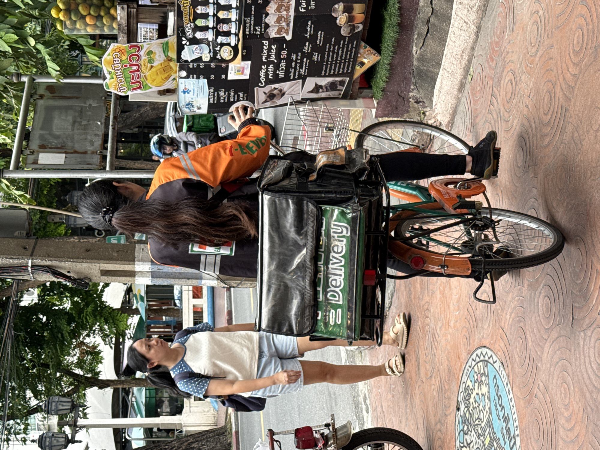 The image depicts a street scene in Bangkok, Thailand. A woman with her hair in a ponytail is riding a bicycle with a large green delivery bag on the back. She is wearing an orange jacket with "7-Eleven" logo and black pants. A woman in a white t-shirt with blue polka dots and denim shorts stands to the left, facing the rider. A menu board is visible to the right, displaying food and drink items with prices in Thai Baht and English. The scene is set on a sidewalk with patterned brickwork, next to a street with trees and buildings in the background. An orange motorcycle is parked on the left side of the image.