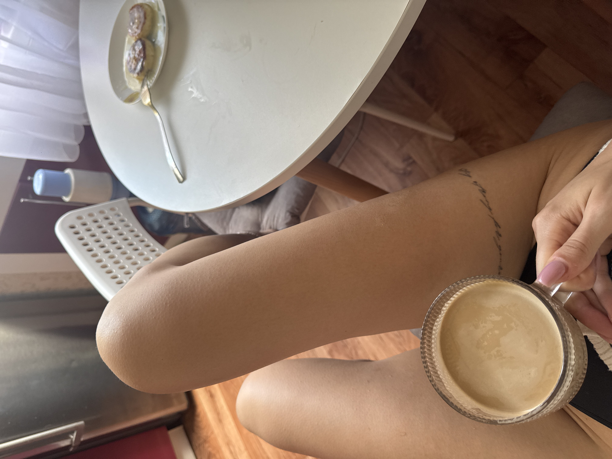 A first-person perspective shot shows a person sitting at a white table, holding a clear glass mug filled with coffee. The mug has a textured rim. In front of the mug, on the table, is a white plate with two small, round pastries. A fork rests on the plate, with one prong inserted into a pastry. To the left of the table, a white chair with a perforated seat is visible, and further left, part of a stainless-steel appliance, possibly a refrigerator, is in the frame. A tall, light blue and white object stands behind the chair, near a sheer white curtain. The floor appears to be made of wood planks. The person has a tattoo on their left thigh. The scene is indoors, suggesting a domestic setting in the city of Chaikovsky, Russia.
