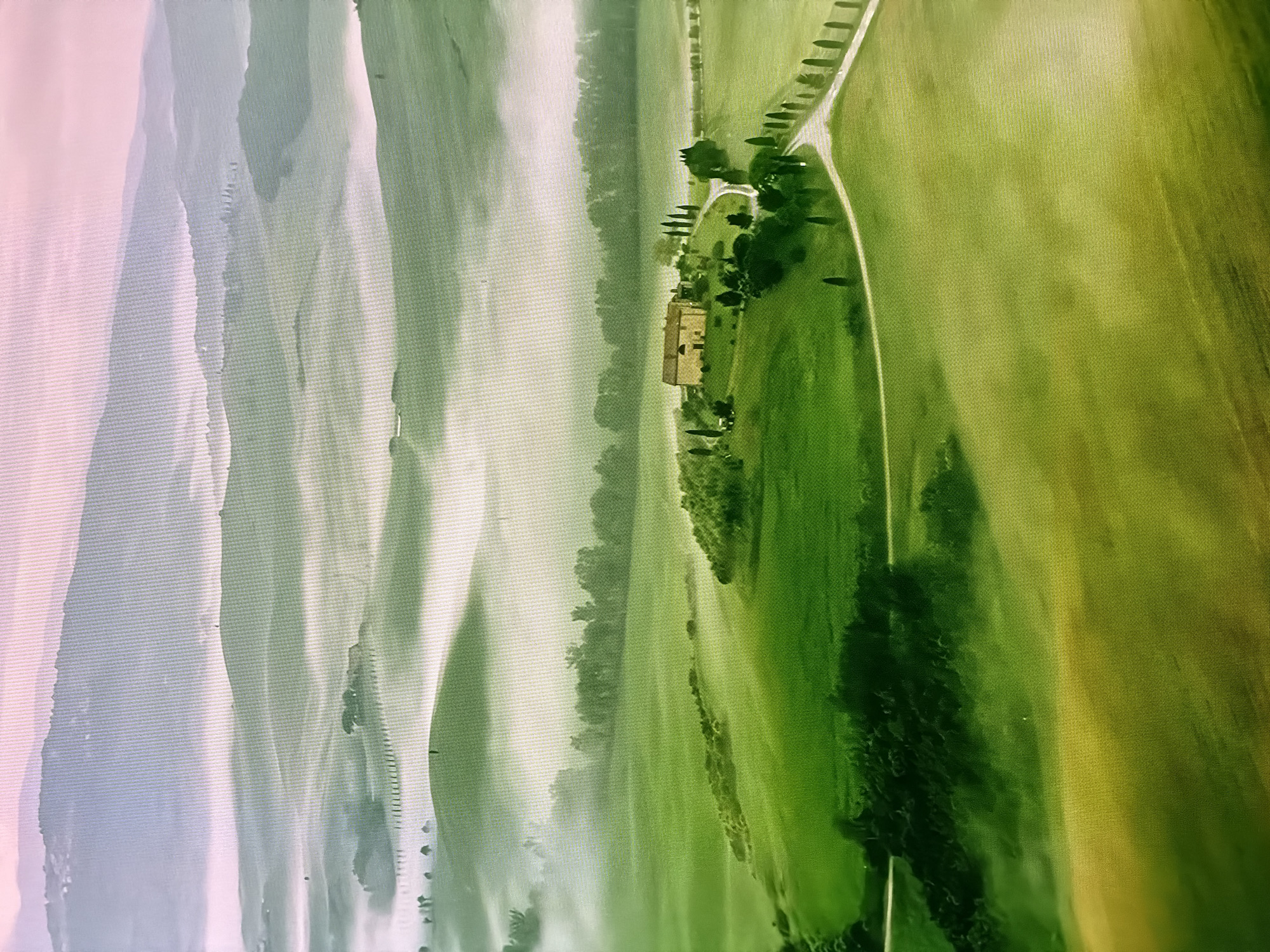 This is an aerial photograph of a rural landscape, likely in Tuscany, Italy, given the rolling green hills, cypress trees, and a solitary farmhouse. The scene is bathed in soft, diffused light, suggesting an early morning or late afternoon setting, possibly with fog or mist settling in the valleys. The foreground features vibrant green fields with gentle slopes. A winding road leads up to a cluster of buildings that appear to be a farmhouse or villa. Tall, slender cypress trees line sections of the road and surround the property, a characteristic feature of the Tuscan countryside. In the midground, more rolling hills are visible, partially obscured by a light mist. The background reveals layered mountains, fading into a hazy, soft pinkish sky, creating a sense of depth and atmosphere. The overall impression is one of tranquility and bucolic beauty. The image does not depict any people, specific landmarks in Chirchik, Uzbekistan, or any discernible actions or interactions.