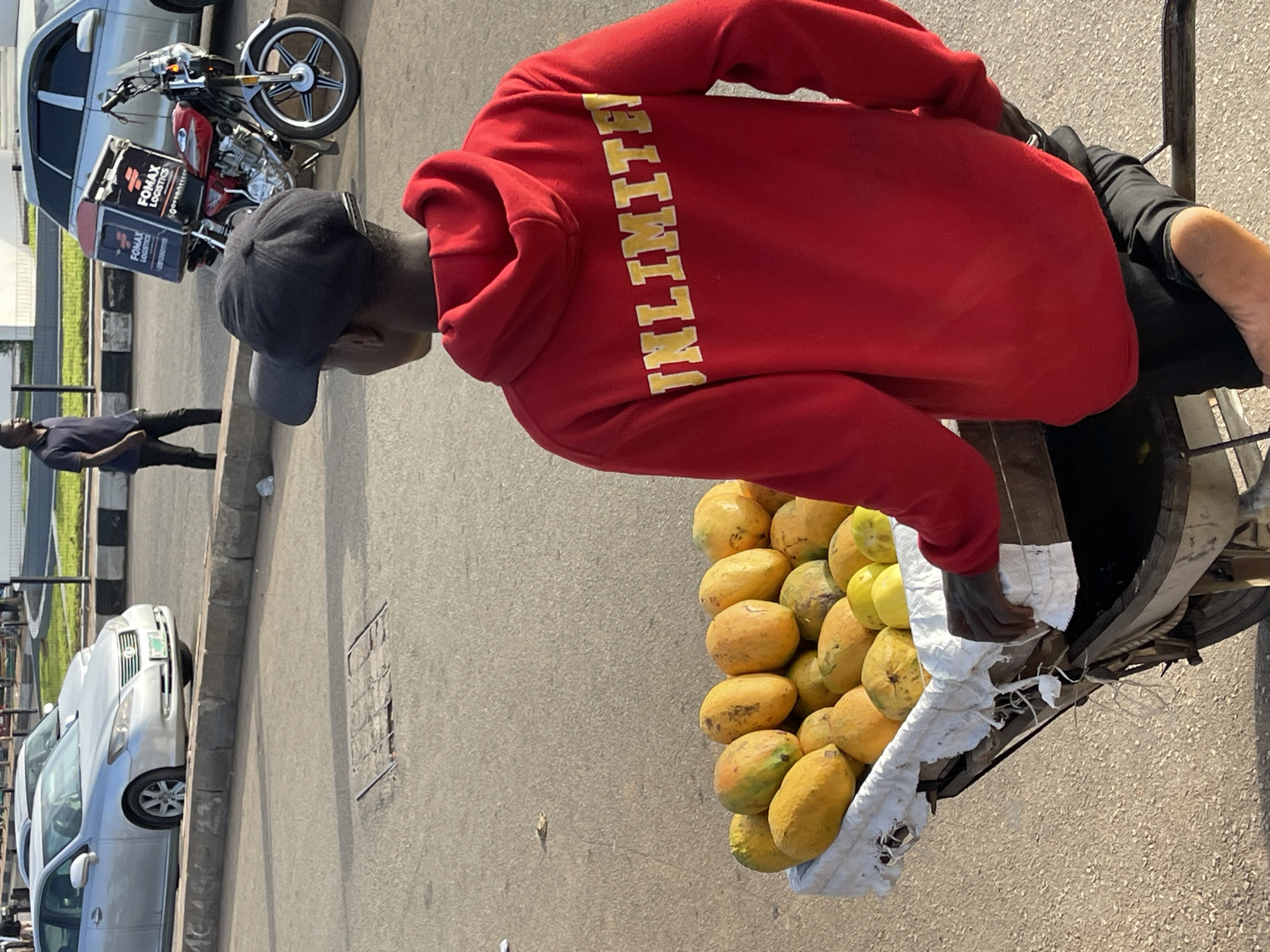 A street scene in Jos, Nigeria is depicted in the photograph. The main subject is a person wearing a red hoodie with the word "UNLIMITED" in yellow letters vertically printed on the back. The person is also wearing a dark cap and is seen from the side, standing next to a cart filled with mangoes. The cart appears to be attached to a bicycle.
In the background, there's a parked motorcycle and a car. A pedestrian walks along the sidewalk. The setting appears to be a paved street with parked vehicles and other urban elements such as sidewalks and curbing. The lighting suggests it's daytime.