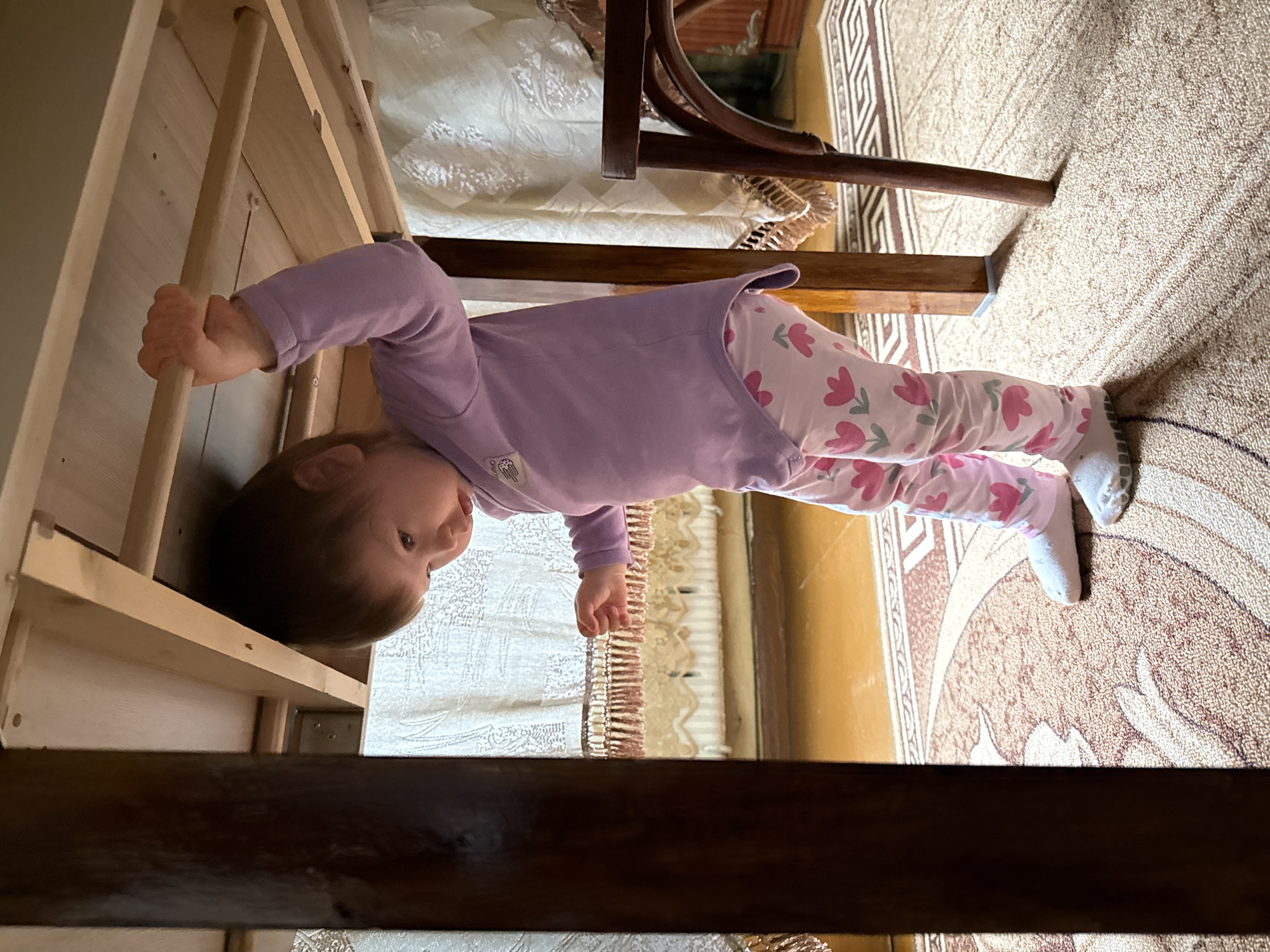 A toddler in a purple long-sleeved shirt and pink floral pants is climbing a wooden structure resembling a ladder. The toddler is facing towards the camera with their head near the top of the ladder. The ladder is constructed from light-colored wood. The toddler is holding on to a rung with one hand. Beneath the ladder is a patterned carpet, and a wooden chair is partially visible in the background. The scene is indoors. There is no verifiable information to indicate this image was taken in Krapivka, Russia.