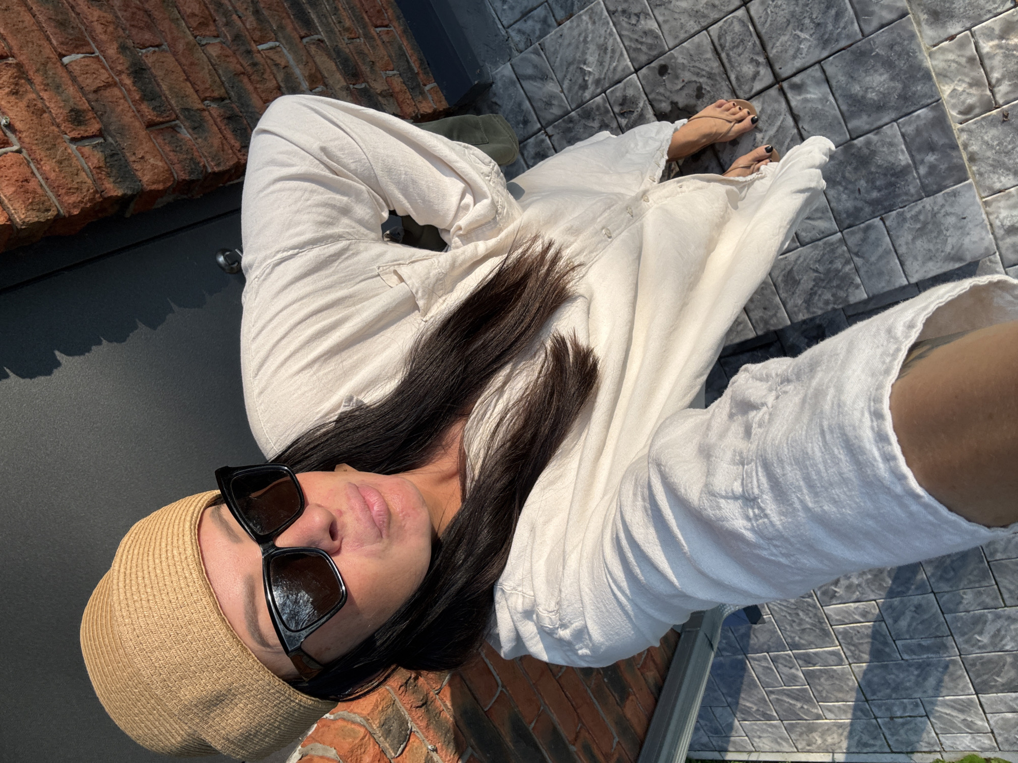 A woman with long dark hair wears a tan straw hat and black sunglasses. She is dressed in a loose, long-sleeved, off-white shirt dress. The woman is standing outdoors on a tiled patio. Behind her, a brick wall and a dark surface are visible. The sunlight casts shadows on the ground. The image appears to have been taken in Chaikovsky, Russia.