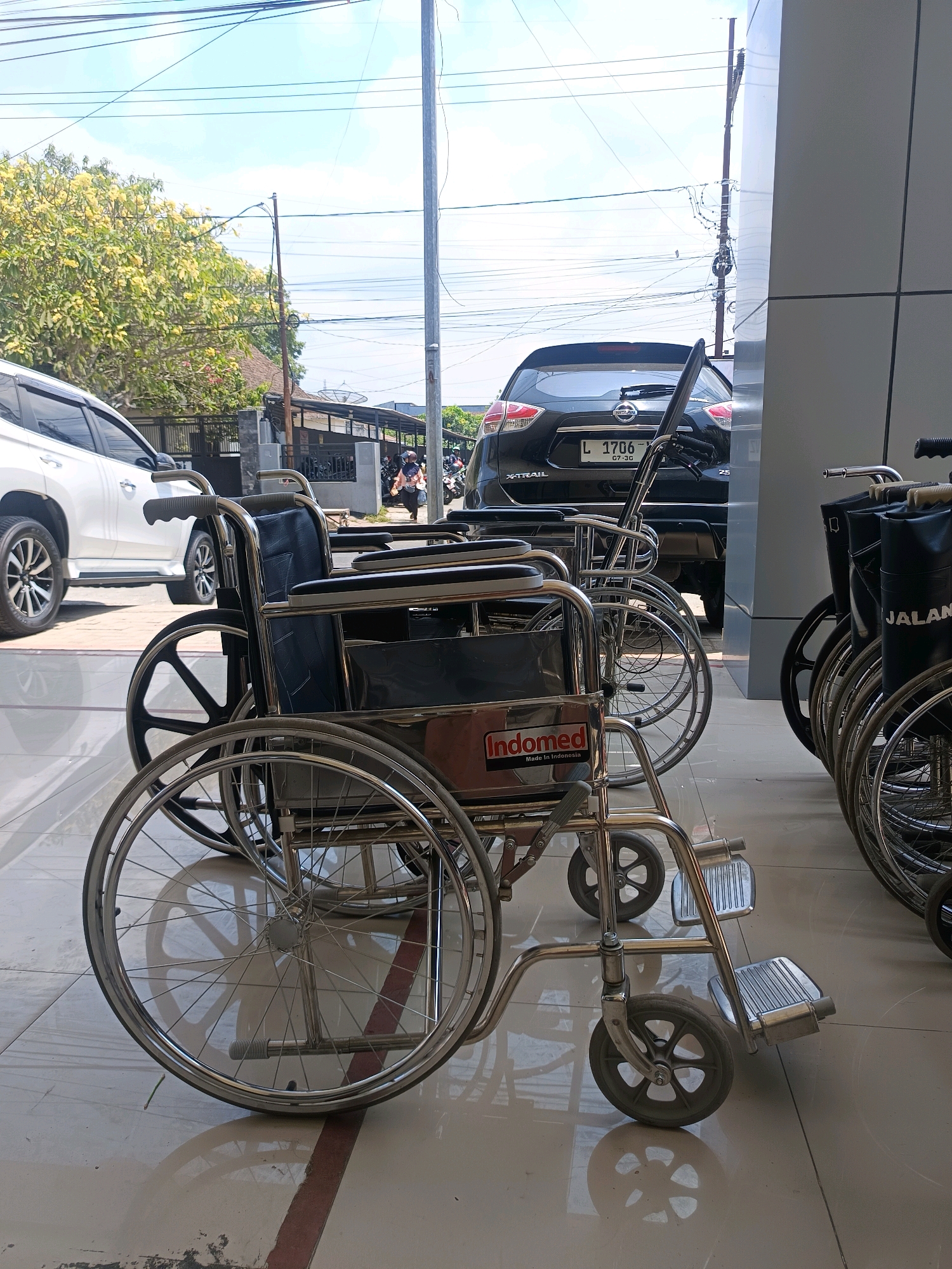 The image captures a row of metal-framed wheelchairs parked on a tiled floor, likely in front of a building. The wheelchairs are made of shiny metal with black upholstered seats and backrests. Some of the wheelchairs have visible logos, including "Indomed" and "JALAN". In the background, a black Nissan X-Trail car with license plate "L 1706" is parked, and a white SUV is also visible. Beyond the vehicles, there are trees with yellow flowers, a fence, and houses, suggesting an urban or suburban setting. Overhead, a network of power lines crisscrosses the sky, which is partly cloudy. The lighting suggests it is daytime, with shadows cast by the wheelchairs and other objects on the ground. The overall scene appears to be a public space, possibly a medical facility or a transportation hub, in Bondowoso, Indonesia, given the license plate format.