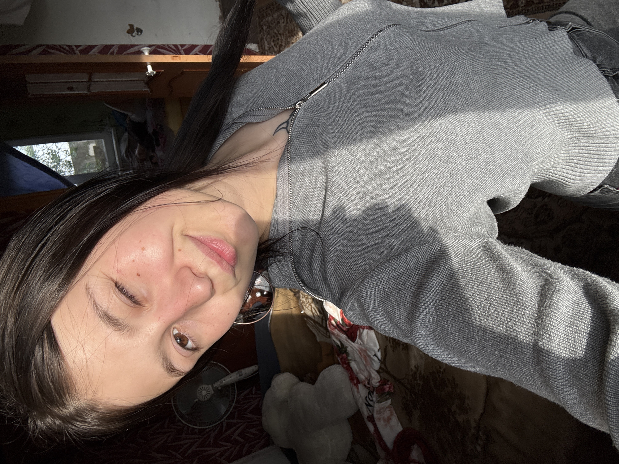 A woman is taking a selfie indoors, likely in a residential setting. She is wearing a gray zippered sweater and her dark hair is parted. One eye is slightly closed in a winking expression, while the other eye is open and brown. Her skin tone appears fair, with visible freckles on her face. She is slightly rotated to the side.
Behind her, a domestic scene is visible with a floor fan and plush toys on a rug. The environment appears to be indoors with a window visible to the top left. No identifying information is present that would suggest the location is Фоки, Russia.