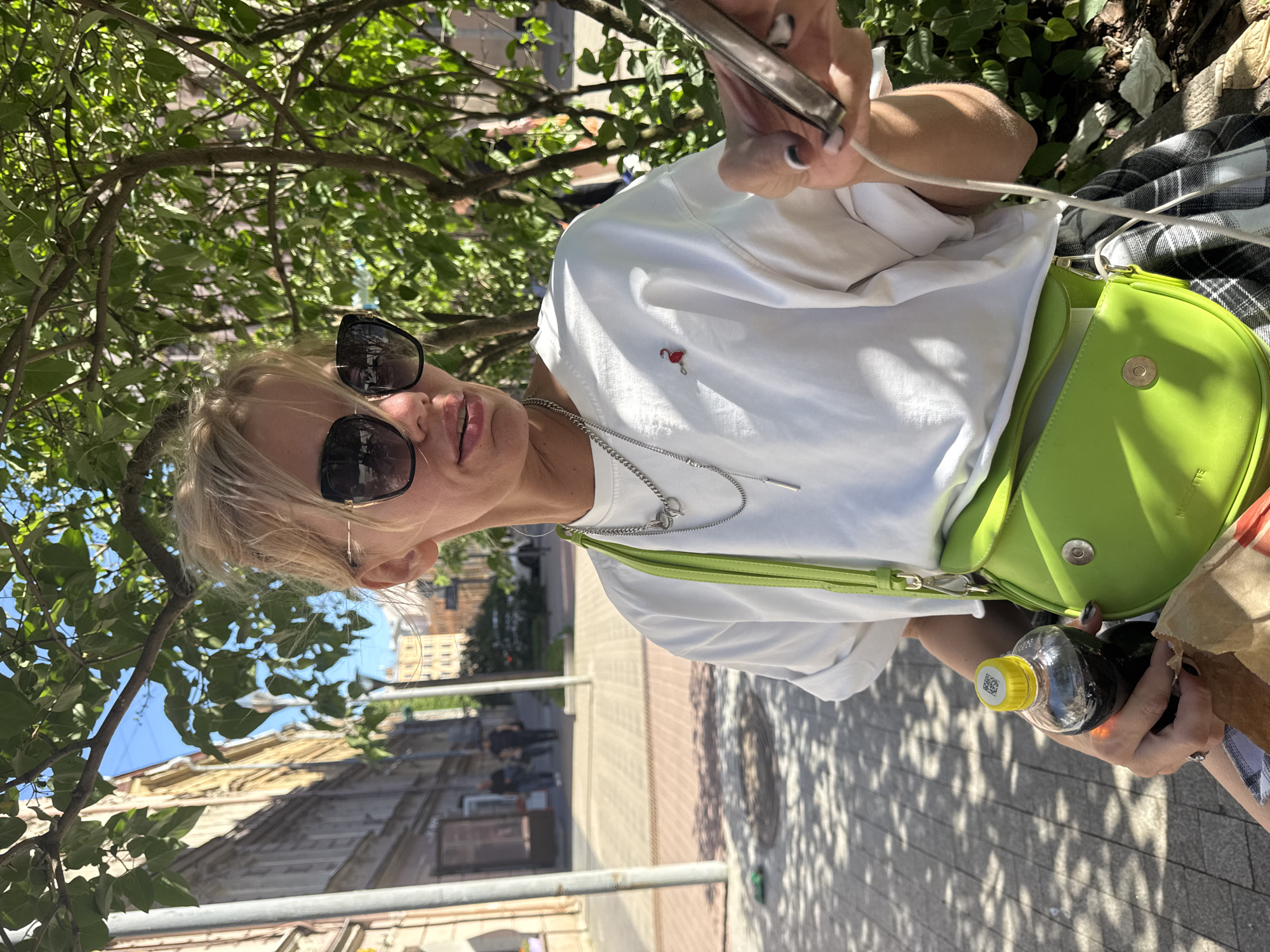 A blonde woman with sunglasses and a white t-shirt is sitting on a bench outdoors, holding a bottle of dark liquid and a phone. She is wearing a lime green cross-body bag and a plaid skirt. The background shows a street scene with buildings and trees. The scene appears to be in Saint Petersburg, Russia, based on the architectural style of the buildings and the cobblestone pavement. The lighting suggests it is daytime, with sunlight filtering through the trees.