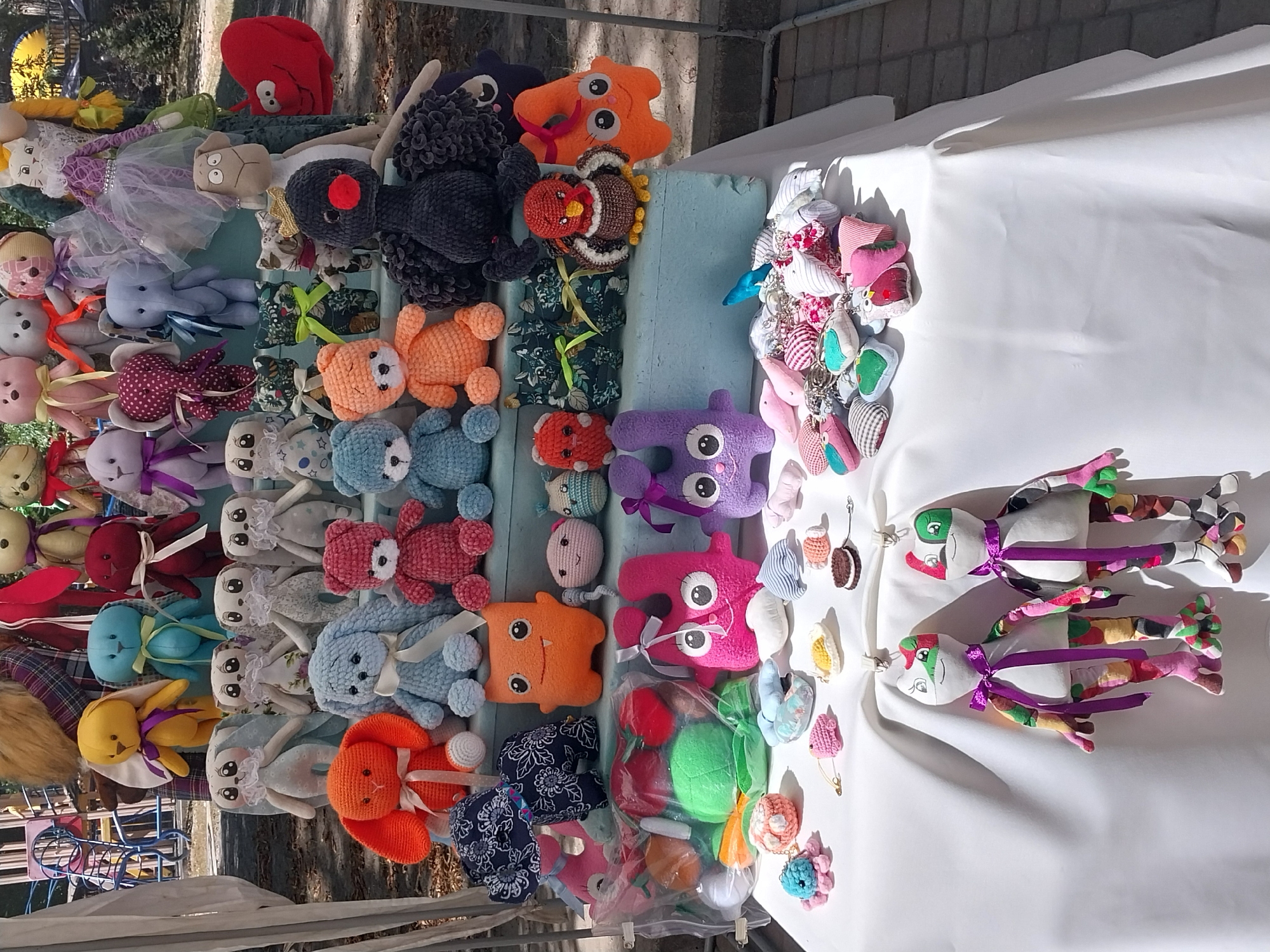 The image depicts an outdoor market stall filled with a variety of handmade stuffed toys. The stall is draped with a white tablecloth, and the toys are arranged on tiered shelves and on the tablecloth itself. The toys include numerous animal figures, such as bears, rabbits, and a turkey, as well as some fantastical creatures. Many of the toys are brightly colored and adorned with ribbons. In the foreground, two fabric figures resembling frogs with patchwork legs are hanging from clips. The background shows a glimpse of a park or playground area with trees and a blue metal structure. The presence of a park setting and the style of the stall suggest a casual outdoor event.