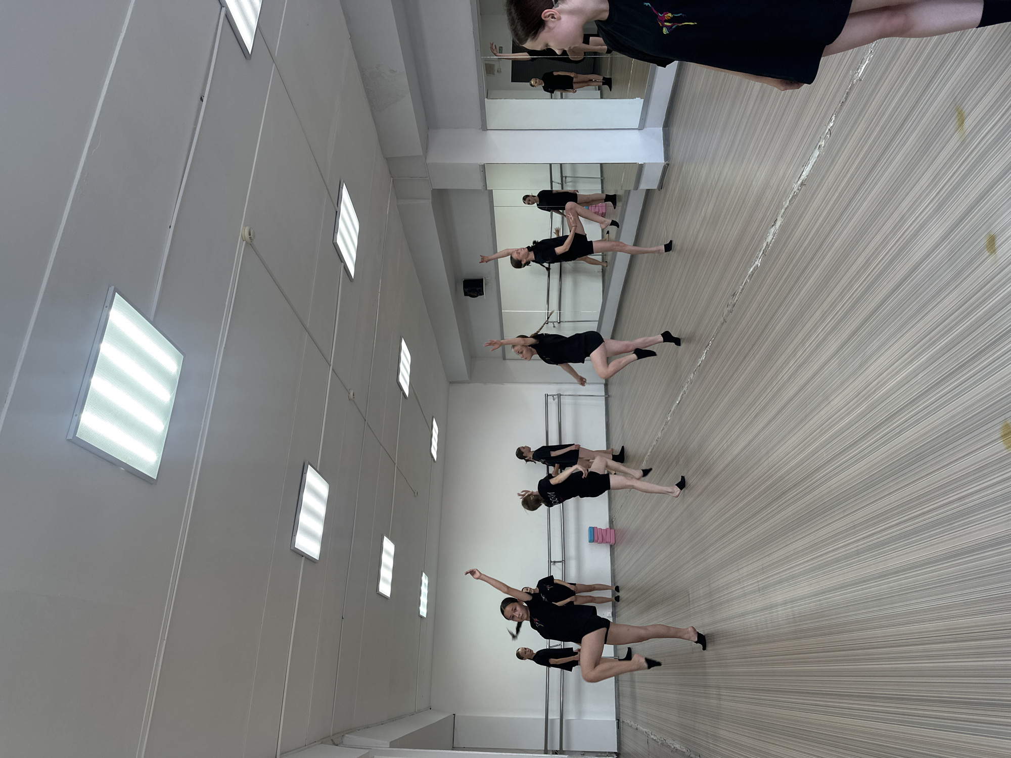 This image depicts a dance studio in Votkinsk, Russia. Several young female dancers, dressed in black leotards and shorts, are practicing ballet. They are positioned throughout the studio, some in mid-pose with outstretched arms and legs, others in the background. The studio features a wooden floor, a mirrored wall with ballet barres, and fluorescent ceiling lights. A stack of pink yoga blocks is visible near the back wall. In the foreground, a young woman in a black t-shirt is partially visible, facing away from the camera. The overall scene captures a dance class in progress.
