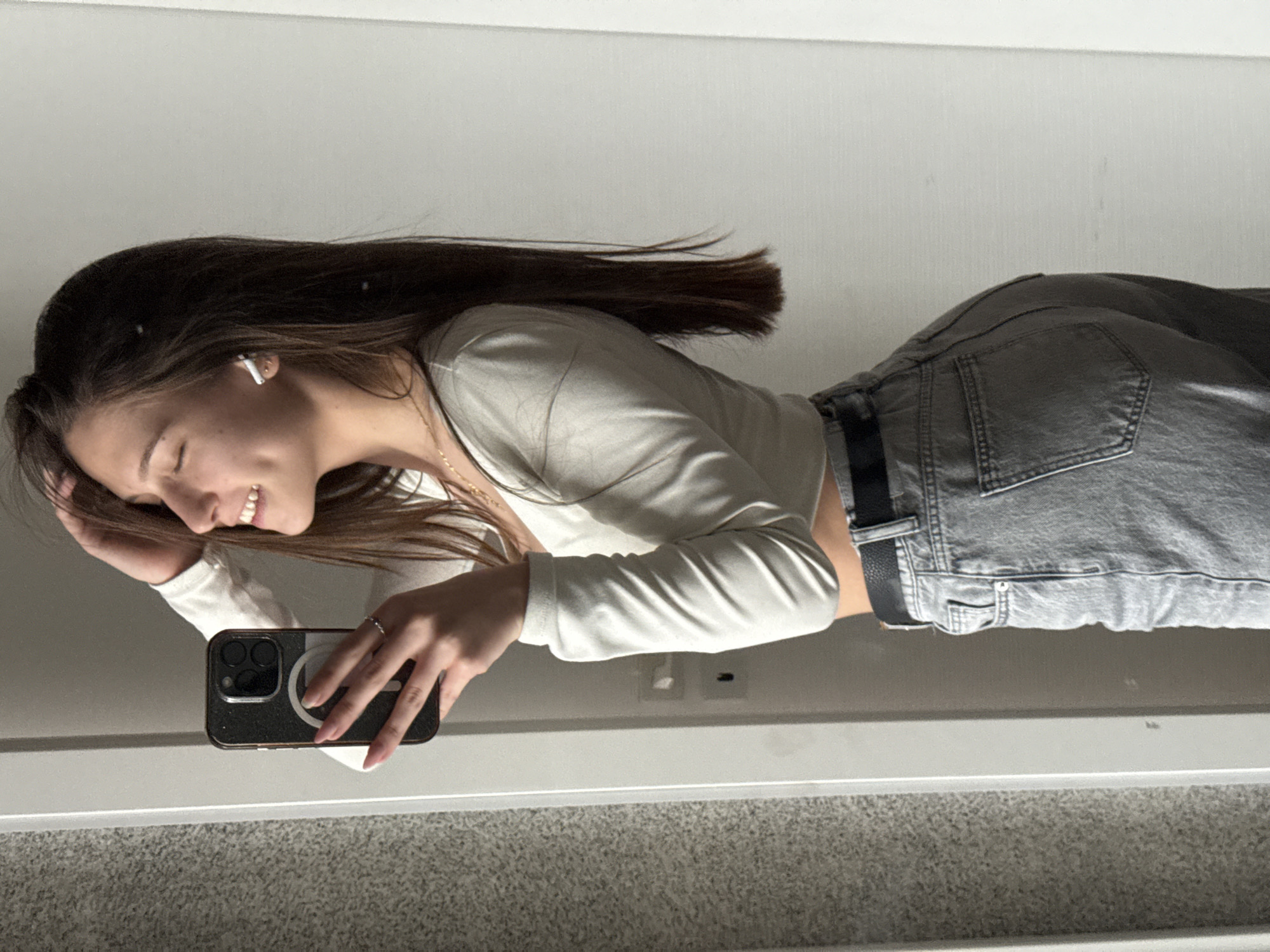 A young woman is taking a selfie in a mirror. She is wearing a white, long-sleeved shirt and grey jeans with a black belt. She has long, dark hair and is smiling slightly. She is holding a dark-colored phone in her left hand. The background appears to be a room with light-colored walls and carpeting. There is no verifiable information suggesting the location is Sofia, Bulgaria.