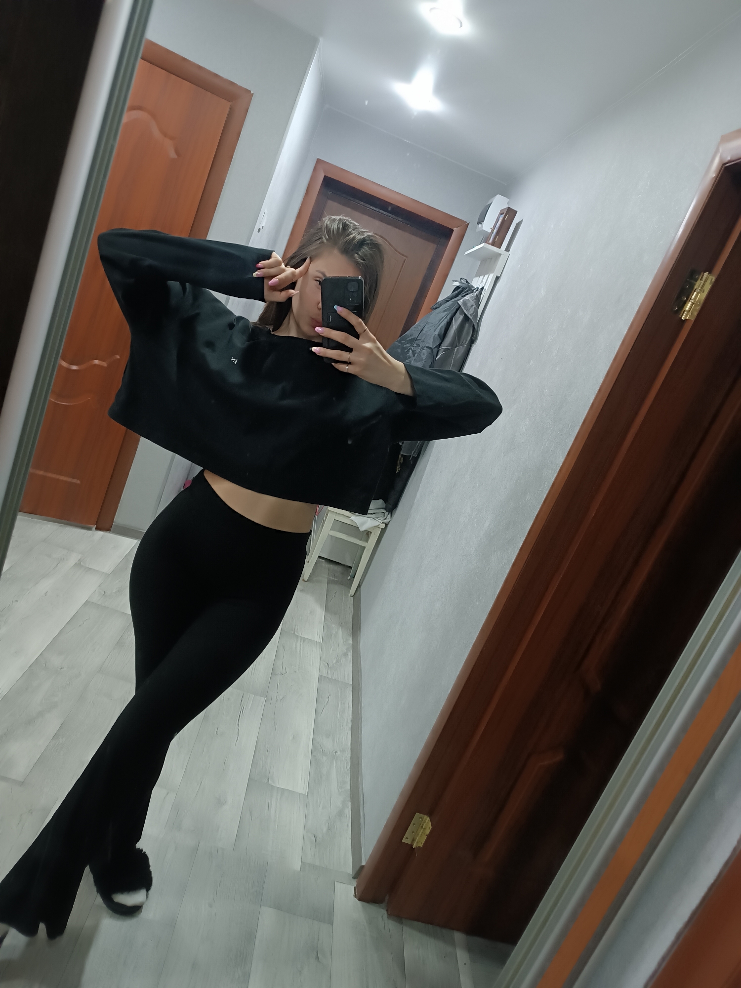 A young woman is taking a selfie in a hallway. She is wearing a black crop top and black leggings, and fuzzy black slippers. She is holding her phone up with both hands. The hallway has wooden doors and light gray walls, with light-colored wood laminate flooring. There is a light fixture visible on the ceiling. There is also a garment on a coat rack. The location is Unknown, Unknown.