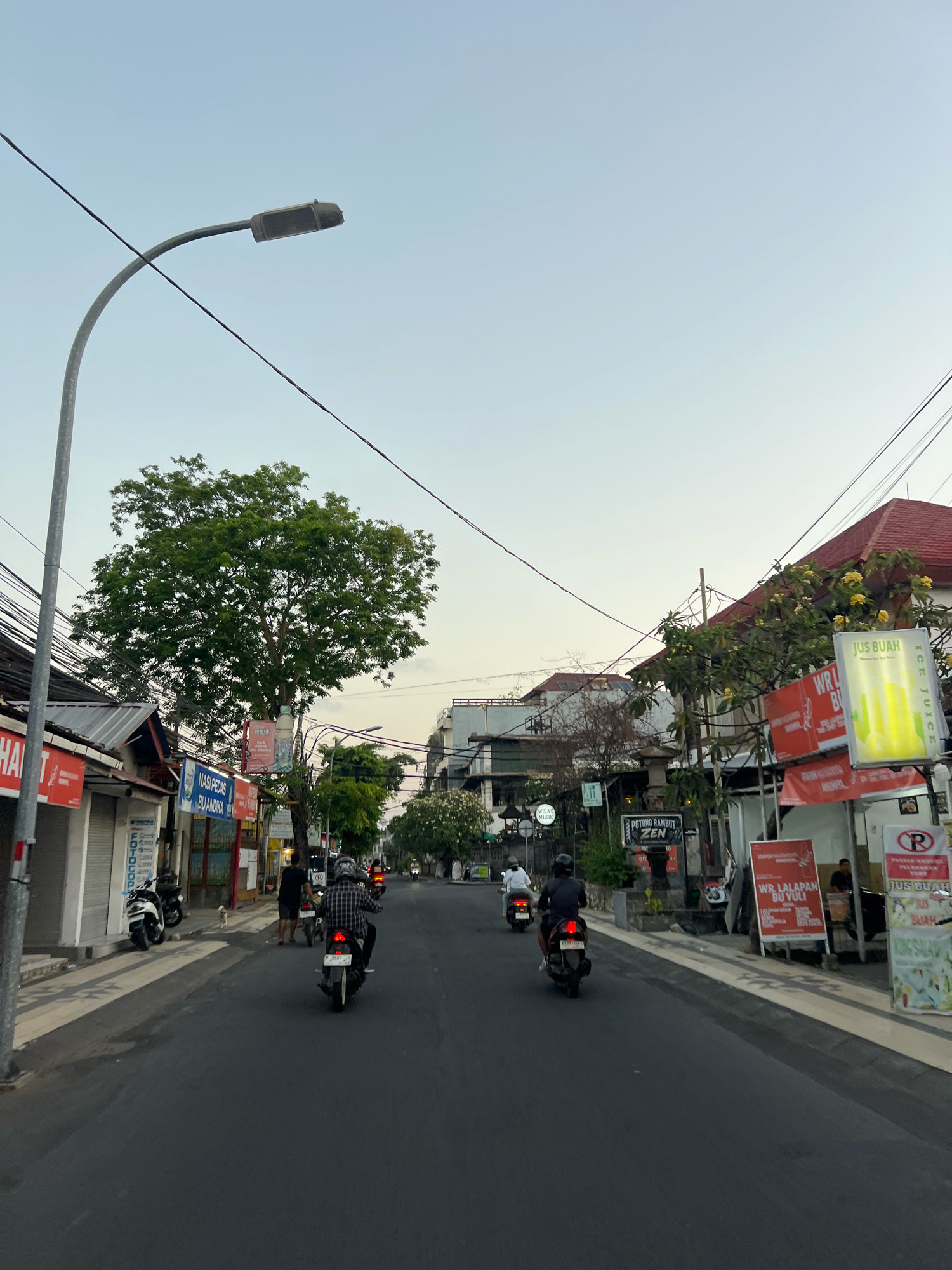 The image depicts a street scene in Kuta, Indonesia, likely during the late afternoon or early evening. The perspective is from the viewpoint of someone on a scooter, looking down a paved road lined with shops and businesses on both sides. Several motorbikes are visible, with three individuals on motorbikes in motion ahead of the viewer, heading away from the camera. One person is wearing a checkered shirt, and another has on a dark helmet and a plaid shirt. A small white dog is also present on the sidewalk.
The buildings flanking the street are commercial establishments, some with closed shutters. Signs in Indonesian are displayed, advertising services such as "Nasi Pedas Bu Andika" (Spicy Rice Bu Andika), "Potong Rambut Zen" (Zen Haircut), and "Jus Buah" (Fruit Juice). A prominent bright yellow sign with green lettering advertises "JUS BUAH ICE JUICE". Trees with green foliage are scattered along the street, with one large, mature tree dominating the left side of the frame. Overhead, a street lamp with a curved arm stands tall, and numerous electrical wires are strung across the sky, a common sight in Indonesian urban environments. The sky is a pale, clear blue, indicating fair weather. The overall atmosphere suggests a typical bustling street in a Southeast Asian city.