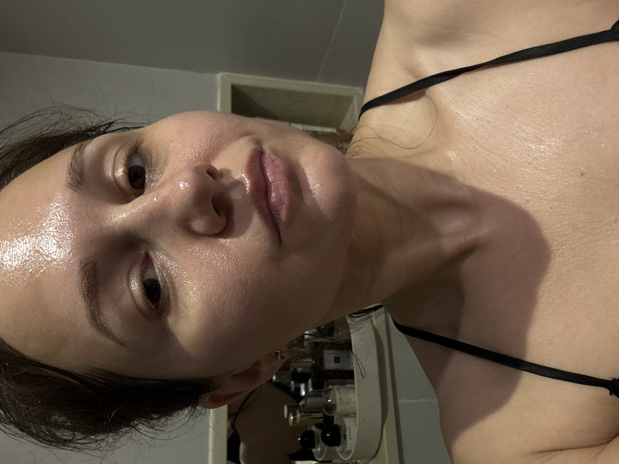 A close-up, sideways-oriented portrait captures a person, likely a woman, with a calm and neutral expression. Her head is tilted upwards and to the left, with her right eye visible, looking towards the upper edge of the frame. Her dark hair frames her face.
The most prominent feature is her skin, which appears highly hydrated, dewy, or wet, glistening under the light. This suggests she might have recently washed her face, applied skincare products, or is perspiring. Her lips are slightly parted, and her eyebrows are well-defined. She is wearing a thin black strap over her right shoulder, likely from a top, bra, or swimwear.
In the blurred background, behind her head, a shelf or counter holds several indistinct bottles and jars, possibly cosmetics or perfumes, with one notable bottle featuring a black top. The setting appears to be an indoor space, such as a bathroom or dressing area, characterized by artificial or dim lighting. There are no clear location cues within the image itself to specifically identify it as Dubai, United Arab Emirates, as provided in the context. No visible text is legible in the image.