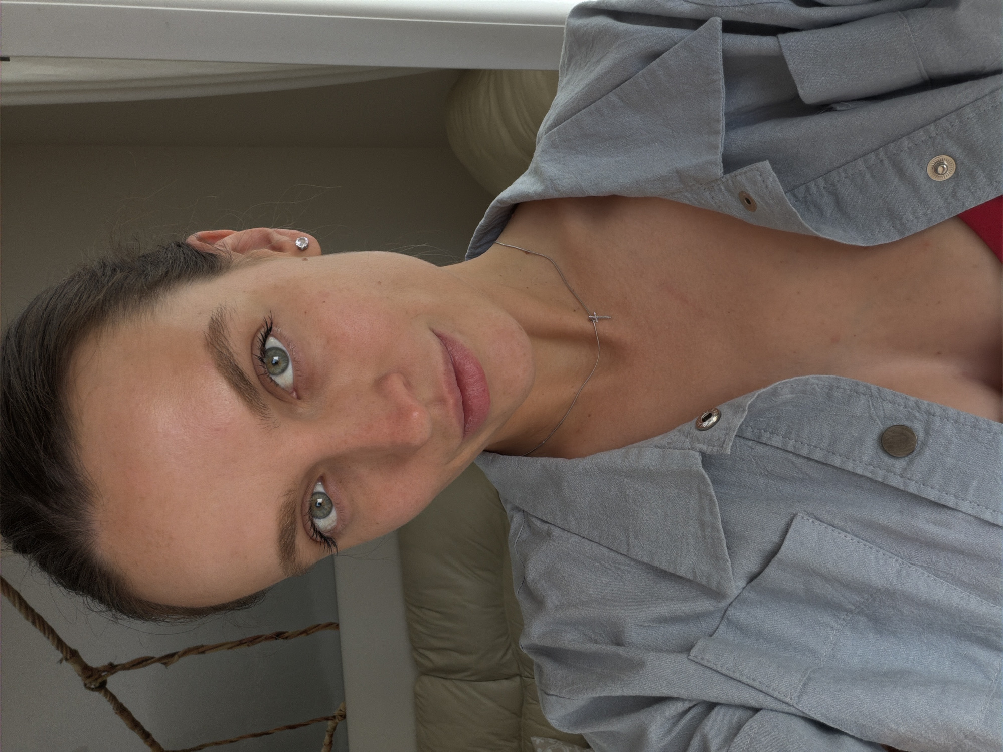 A woman with fair skin, gray-green eyes, and dark hair pulled back is depicted in an indoor setting. She wears a light gray button-down shirt, with glimpses of a red garment underneath. A delicate silver necklace with a cross pendant rests on her chest, and a small stud earring adorns her left ear. The background includes a beige tufted headboard, a rope object, and a white wall. There is no information about Budva, Montenegro.