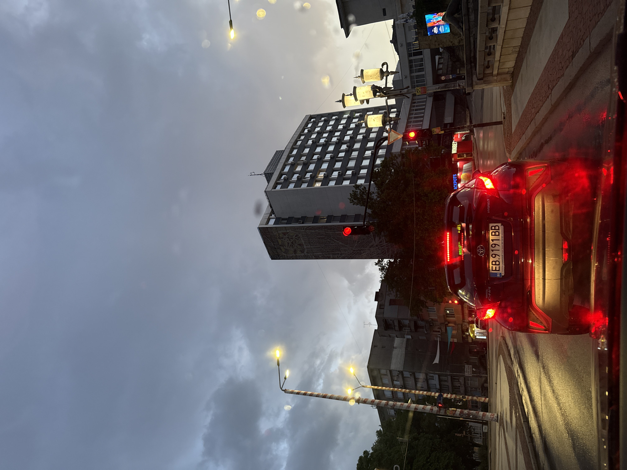 The image captures an urban street scene in Gabrovo, Bulgaria, likely during dusk or an overcast, wet day, given the illuminated lights and reflective wet asphalt. The sky is a dominant grey with heavy clouds, suggesting recent rain or impending downpour, though brighter patches are visible towards the top right.
In the immediate foreground, the rear of a dark-colored Toyota C-HR car is prominently visible, its bright red taillights glowing. The Bulgarian license plate "EB 9191 BB" is clearly displayed on the vehicle. The car is stopped on a wet road, possibly at a red traffic light which is visible further down the street to the left.
The street is lined with buildings. A prominent, multi-story building with a grid pattern of windows and air conditioning units covers a large section of the left frame. Its lower section features a large, detailed mural or architectural design. Scattered streetlights emit a warm yellow light, contrasting with the cool, grey tones of the sky and buildings.
On the right side, other buildings and structures are visible, some featuring illuminated commercial signs. Identifiable text on these signs includes "JACKPOTS," "ROULETTE," and a "Hyundai" logo. Another sign has Cyrillic text that appears to be "ЛЕТИН." The overall impression is a bustling urban environment under moody weather conditions.